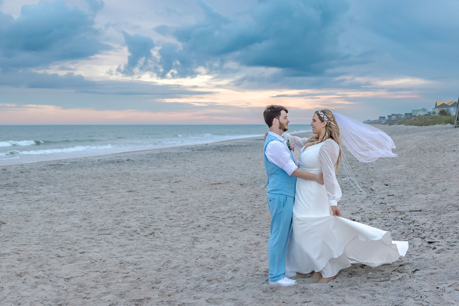 The bride and groom stand together on the sandy beach at sunset with soft pastel skies and gentle waves in the background, captured during a Topsail Beach, NC wedding, blending coastal charm and romantic elegance.