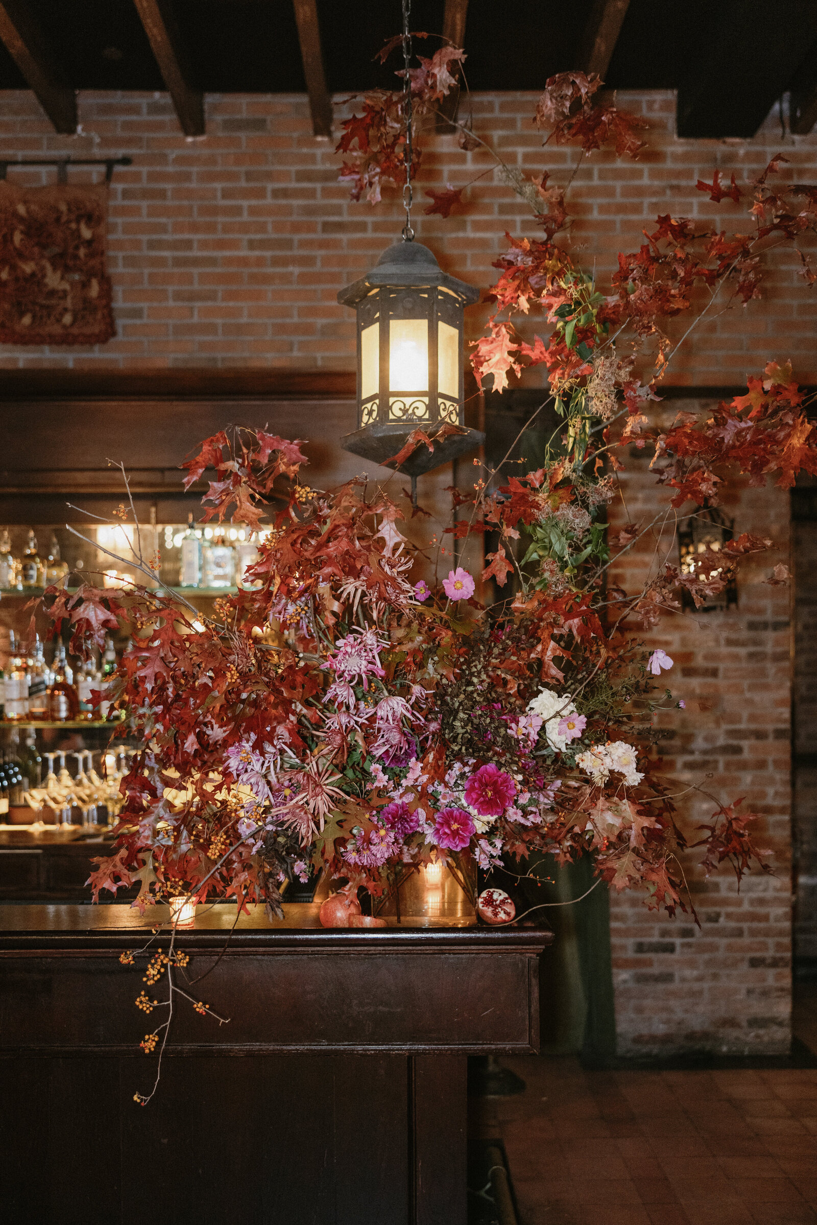 Direct flash photo of fall colored wedding floral installation with candles and glowing lantern at corner of bar in front of brick wall at The Bowery Hotel in NYC