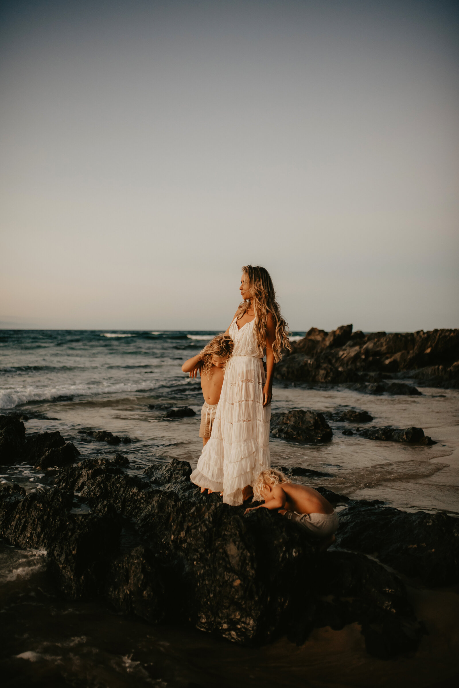 A woman in a white dress stands on a rocky shore, holding the hand of a small child. Another child is nearby, looking at the rocks. The ocean and horizon are in the background.