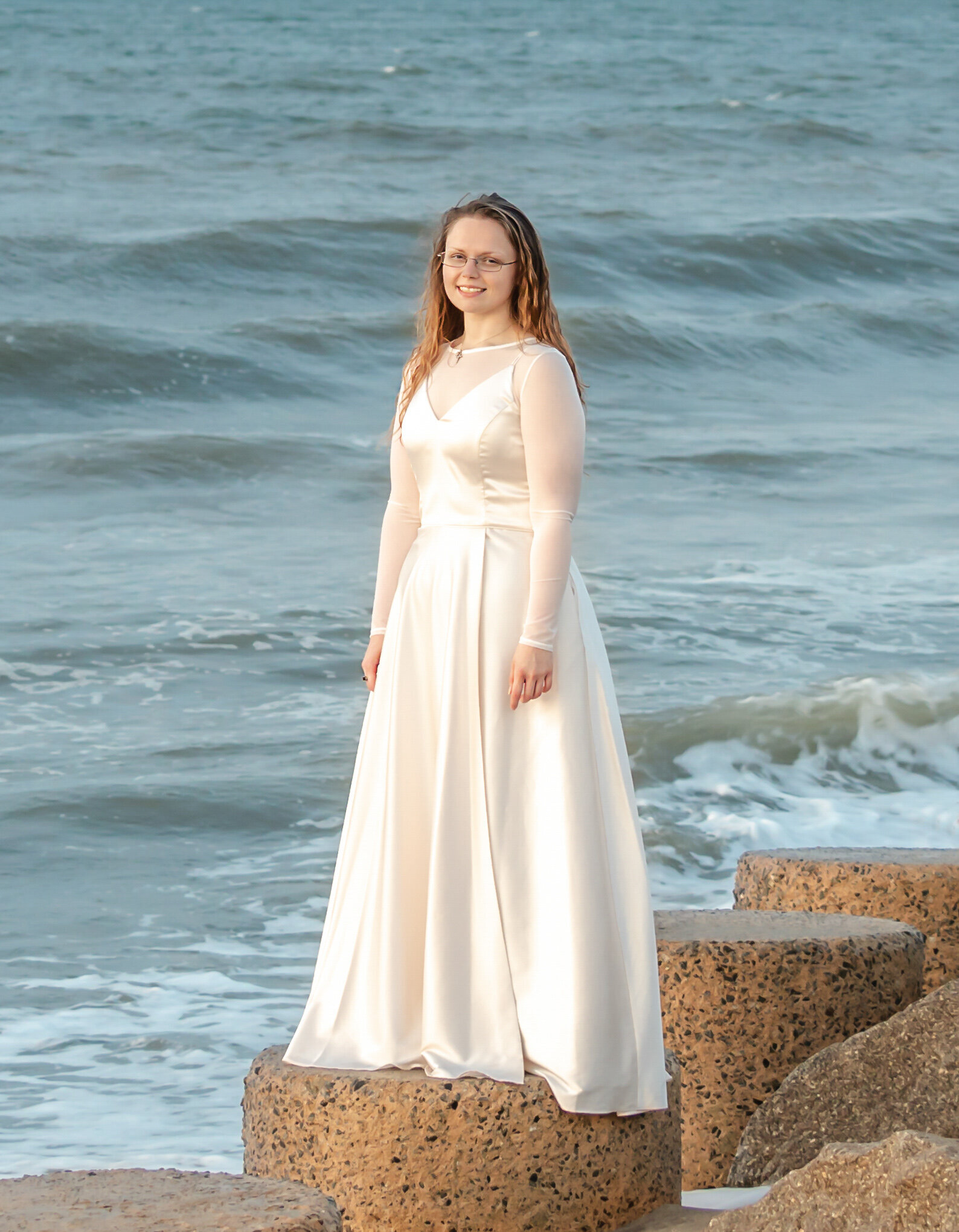 A college senior in a flowing white dress standing on rocks with ocean waves in the background in Wilmington, North Carolina. This serene image captures the elegance of senior photography by the sea, perfect for artistic portraits.