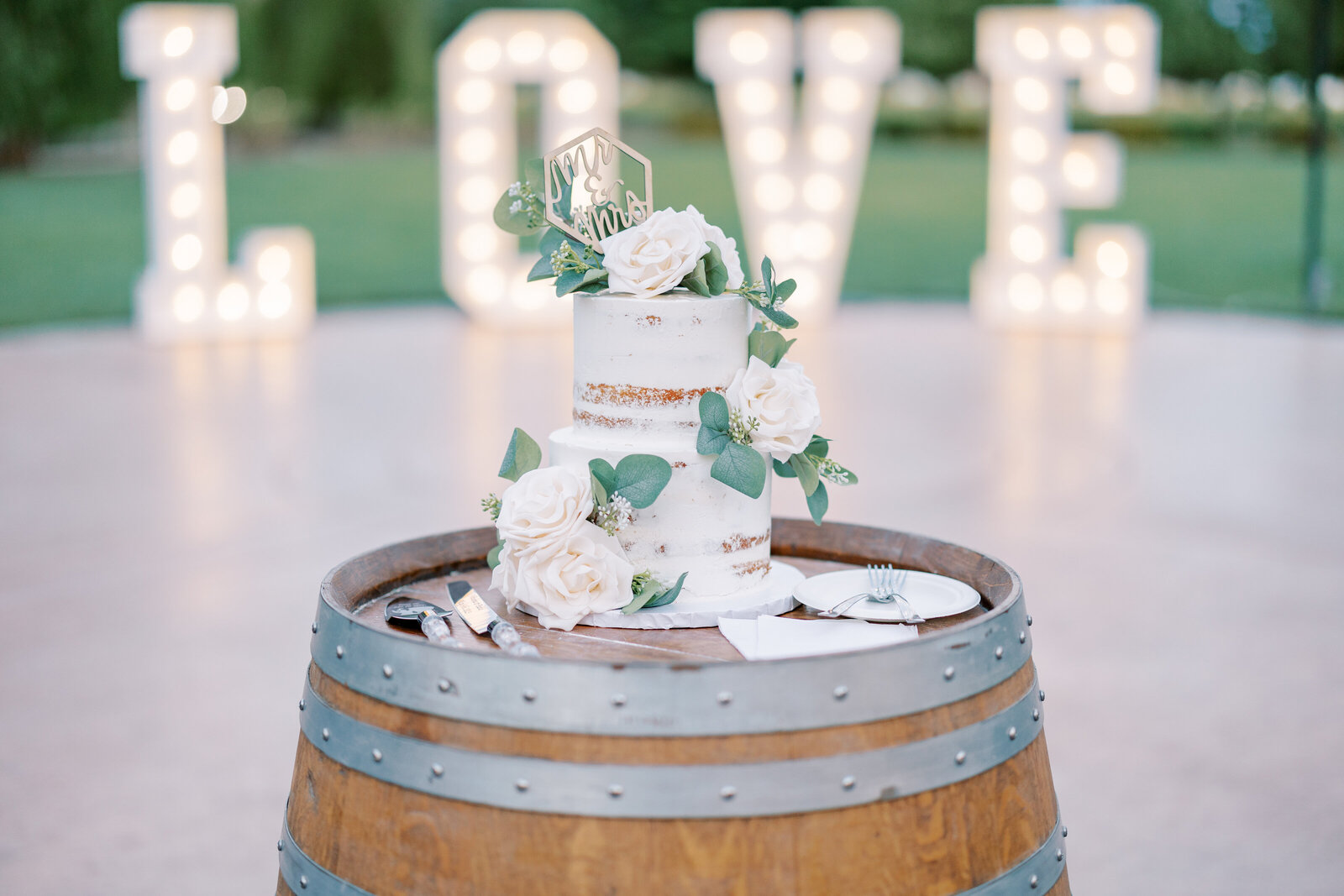 sacramento wedding photographer captures wedding cake with floral decorating the tiers with a cursive name cake topper while sitting on a wood barrel with a light up large letter sign behind the cake