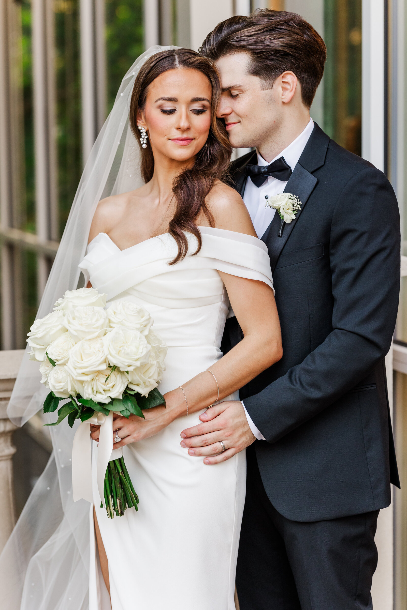 A groom gently embraces his bride holding a bouquet of white roses, both dressed in elegant wedding attire outside a Dallas wedding venue.