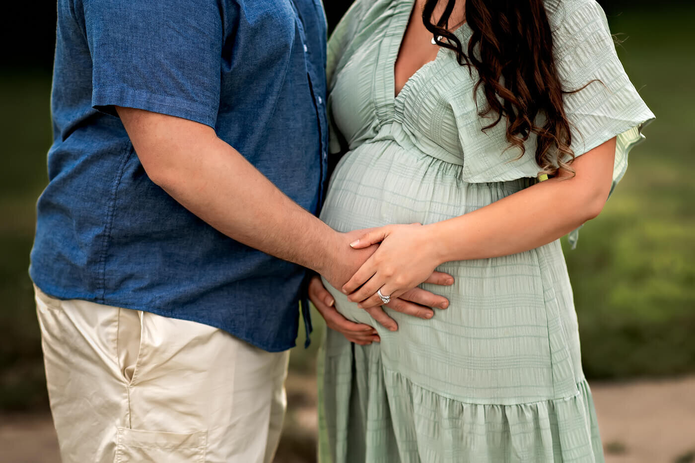 A couple stands close together outdoors, gently holding hands over the woman's pregnant belly—a sweet moment captured in maternity portraits. The woman wears a light green dress, and the man wears a blue shirt and light shorts. Their faces are not visible.