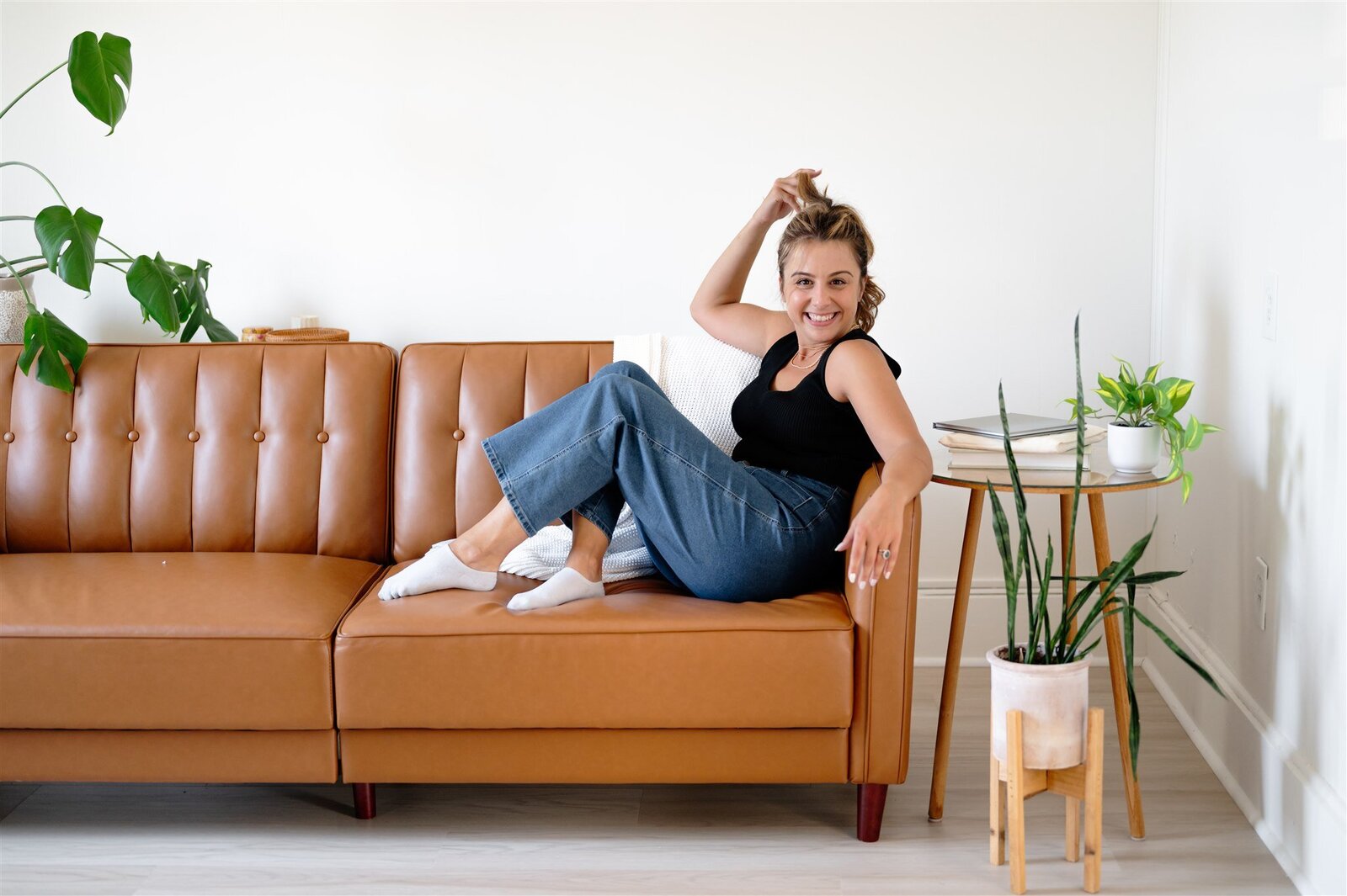 Bee, owner of BusyBSocial, is smiling off to the side with a glass of champagne in her hand as she sits on the couch