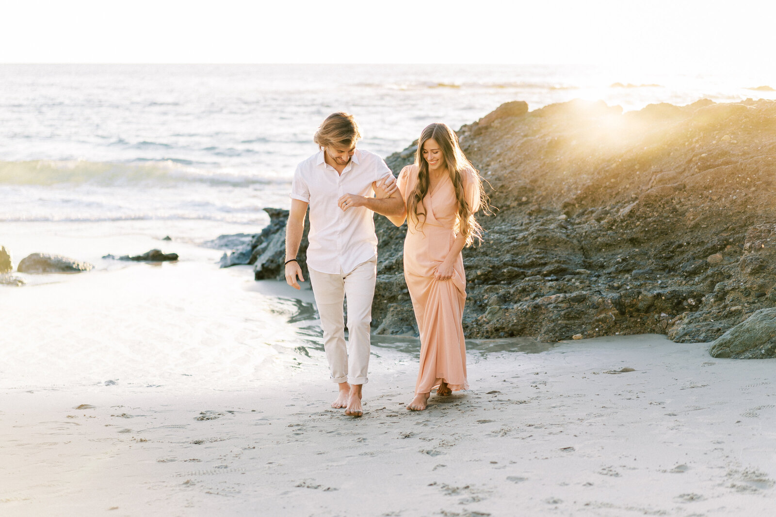 Portrait of a man in a white shirt and khakis locked in arm with a woman in a pink dress on the sand at the beach.