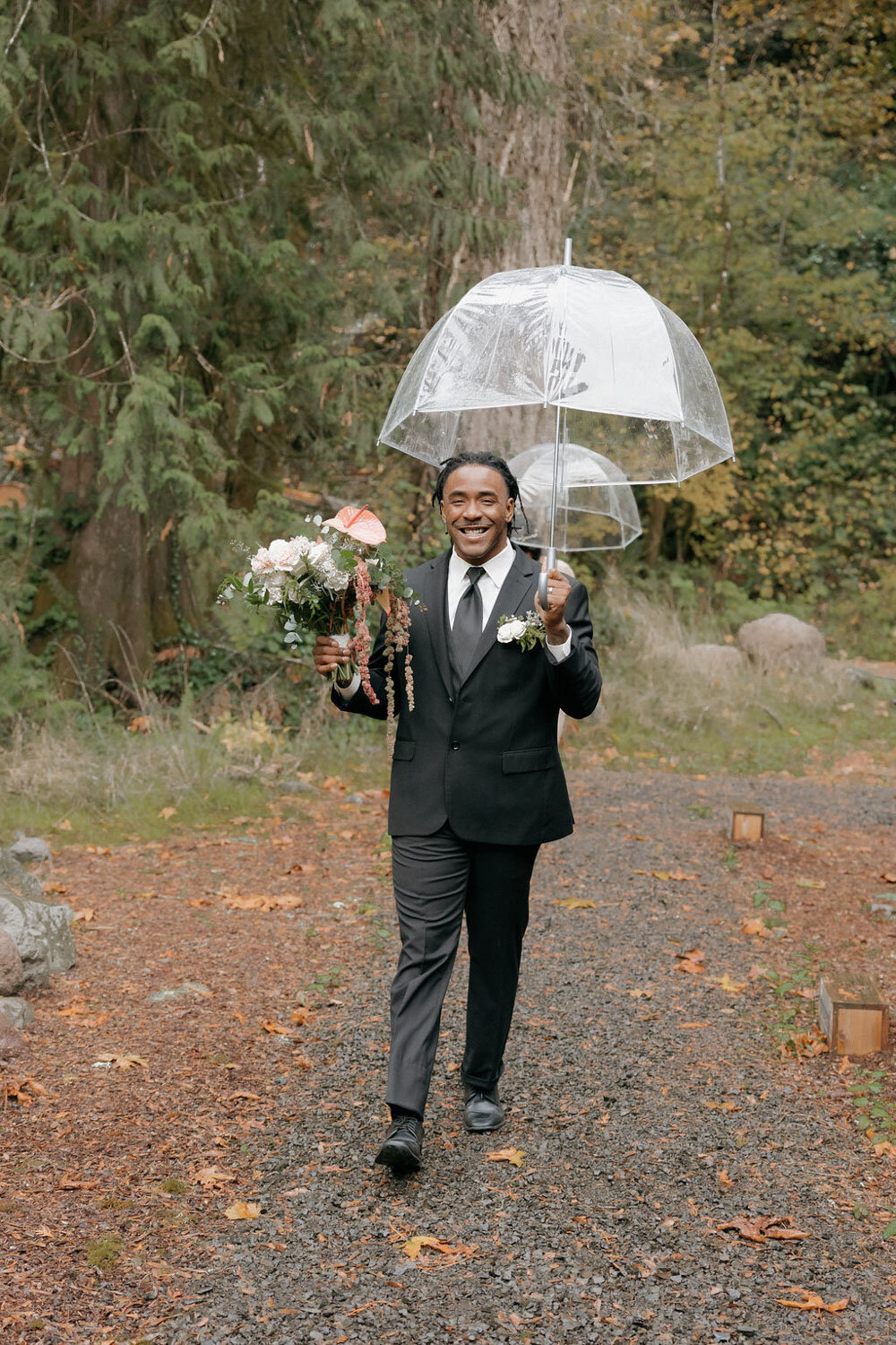 Groom walking down a path holding clear umbrellas and flowers