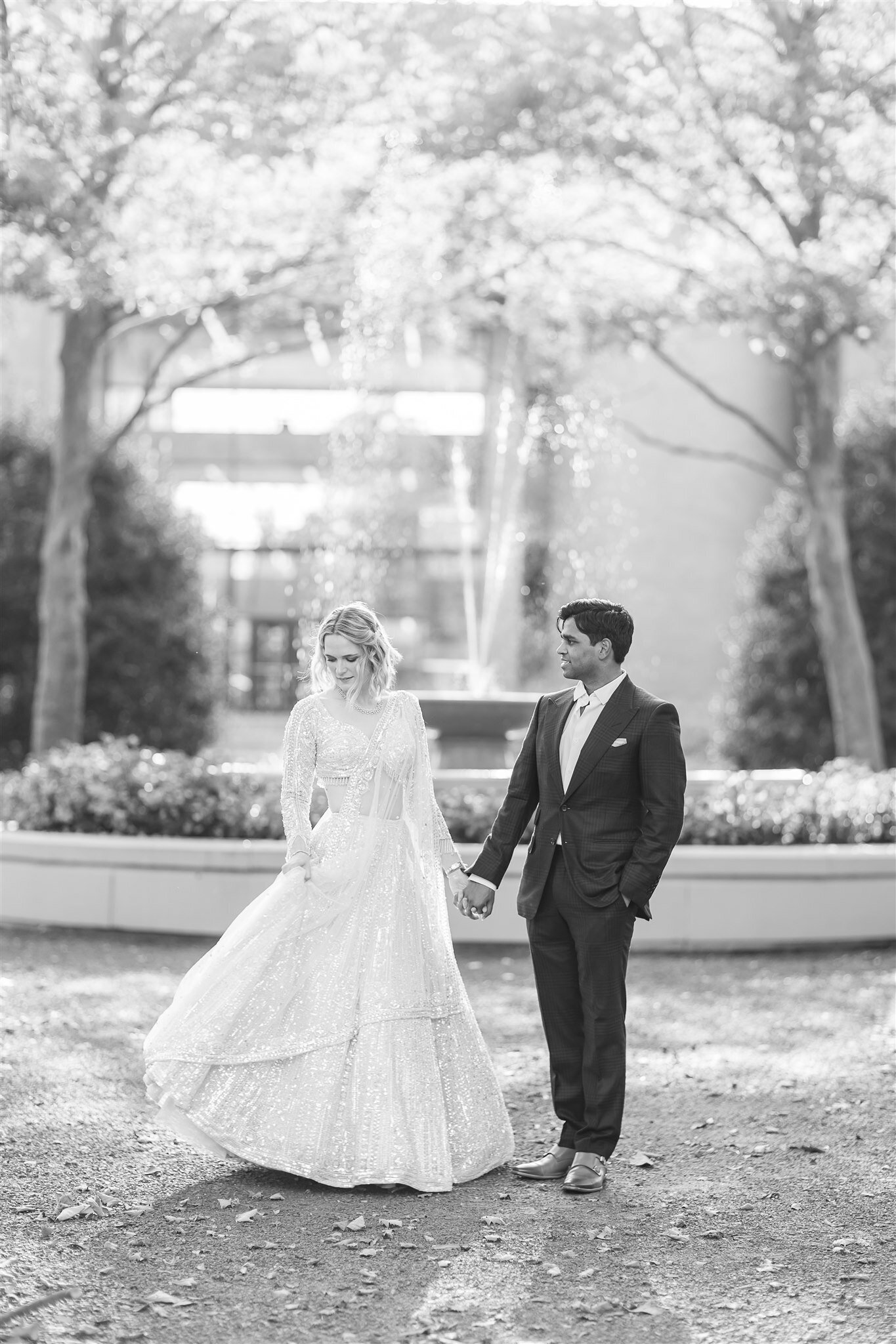 Black and white wedding portrait of a bride in a long-sleeved gown and groom in a classic suit, holding hands near a fountain.
