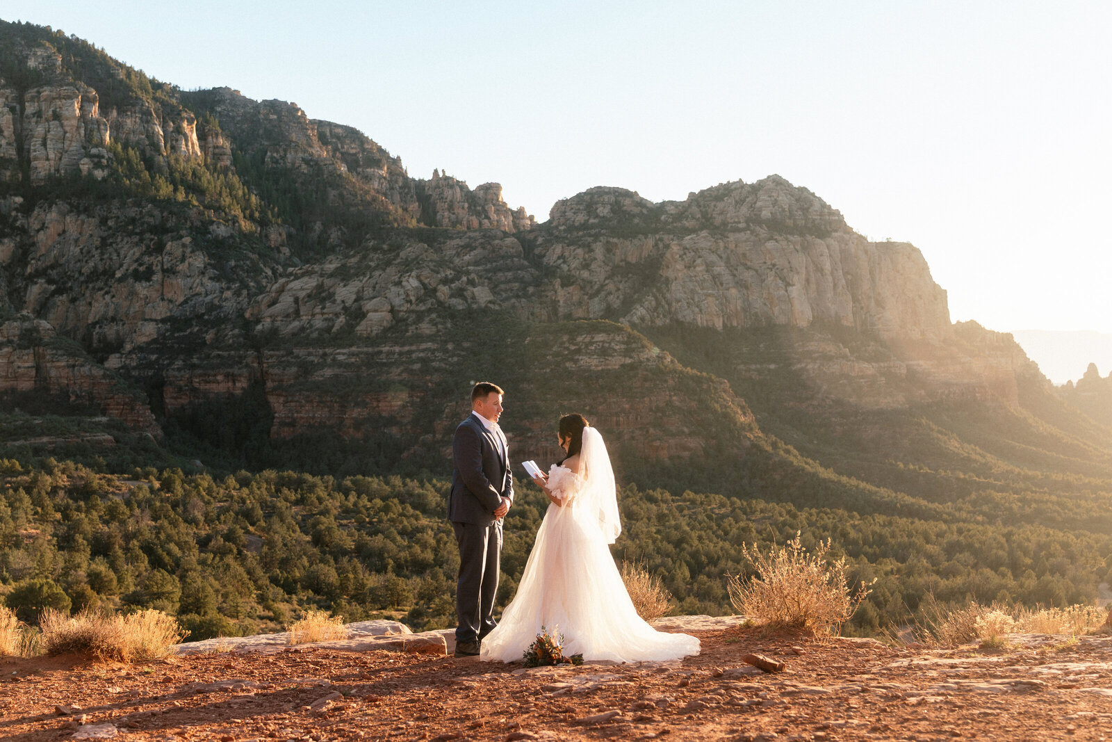 Bride and groom exchanging vows at sunset on the edge of Merry Go Round Rock in Sedona, Arizona, with dramatic mountain views and golden light illuminating the red rock landscape.