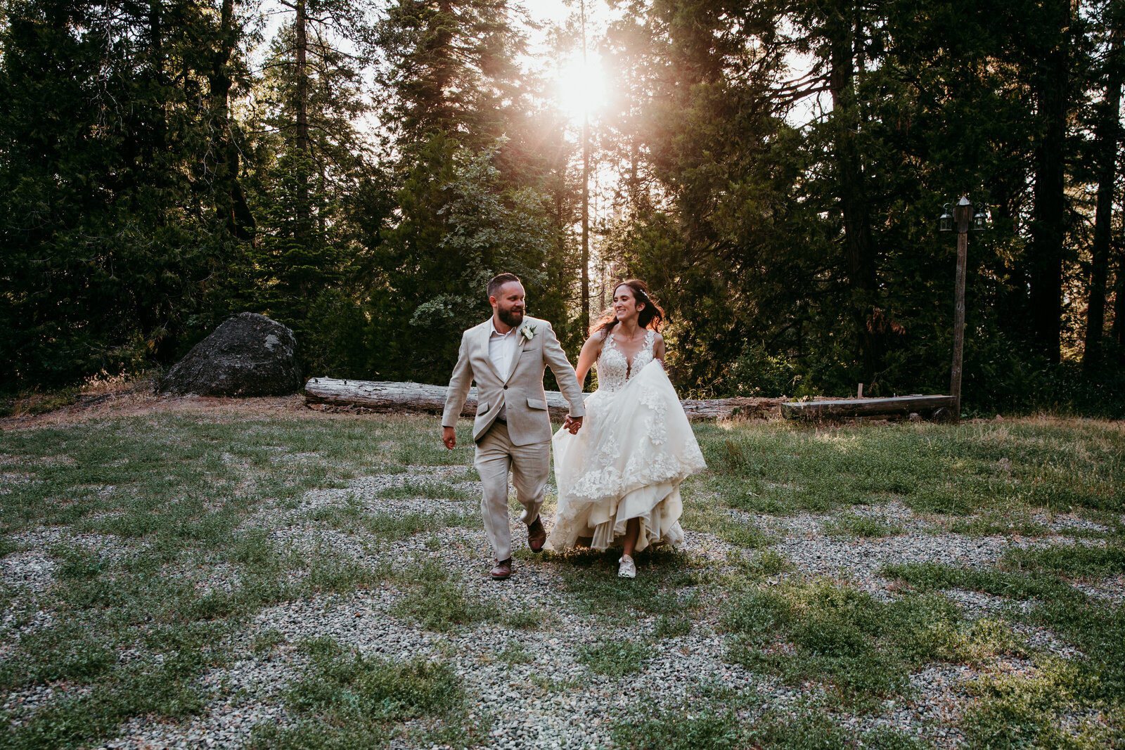 A newlywed couple joyfully runs hand in hand through a sunlit forest clearing near Yosemite, the bride lifting her dress as they embrace the moment