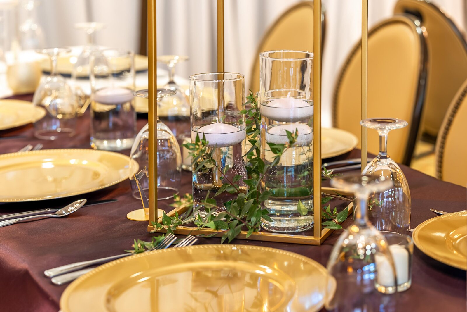A wedding reception table decorated with gold plates, floating candles in glass vases, and greenery inside a gold frame centerpiece, captured during a Boone, NC wedding, showcasing modern elegance.