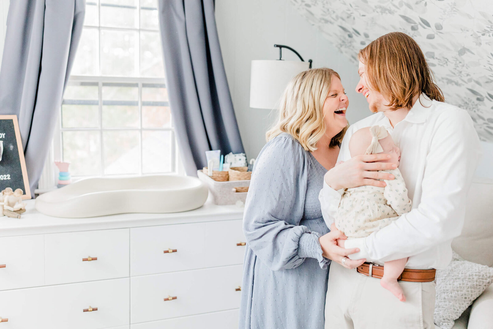 Mom and dad laugh together in the nursery while dad holds their newborn