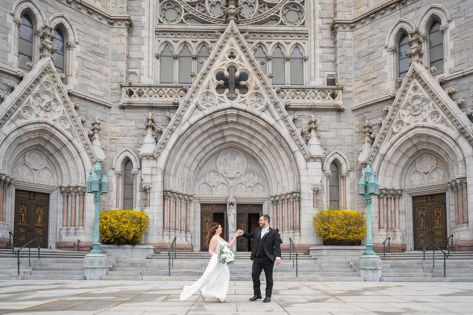 (direct upload)
A bride and groom stand holding hands in front of an ornate stone church with arched doorways and symmetrical architecture.