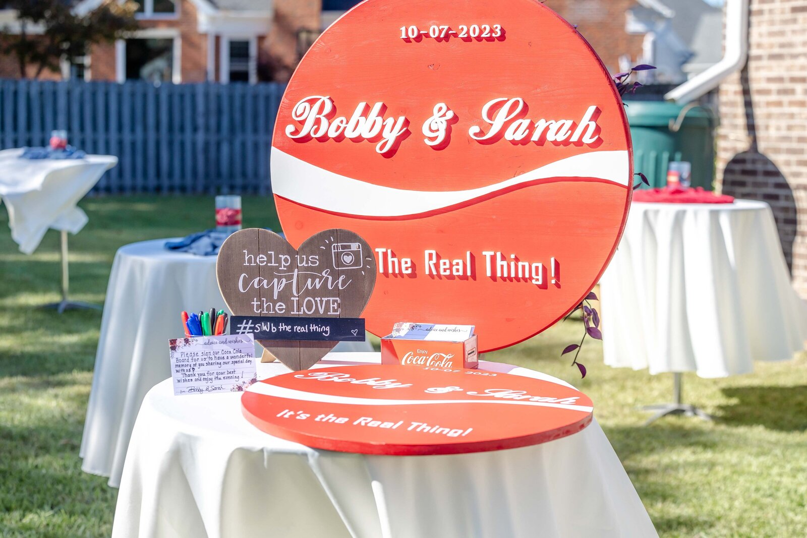 A Coca-Cola-themed wedding sign with "Bobby & Sarah" and guestbook station set up outdoors, captured during a Wilmington, NC wedding, blending unique branding with personalized wedding decor.