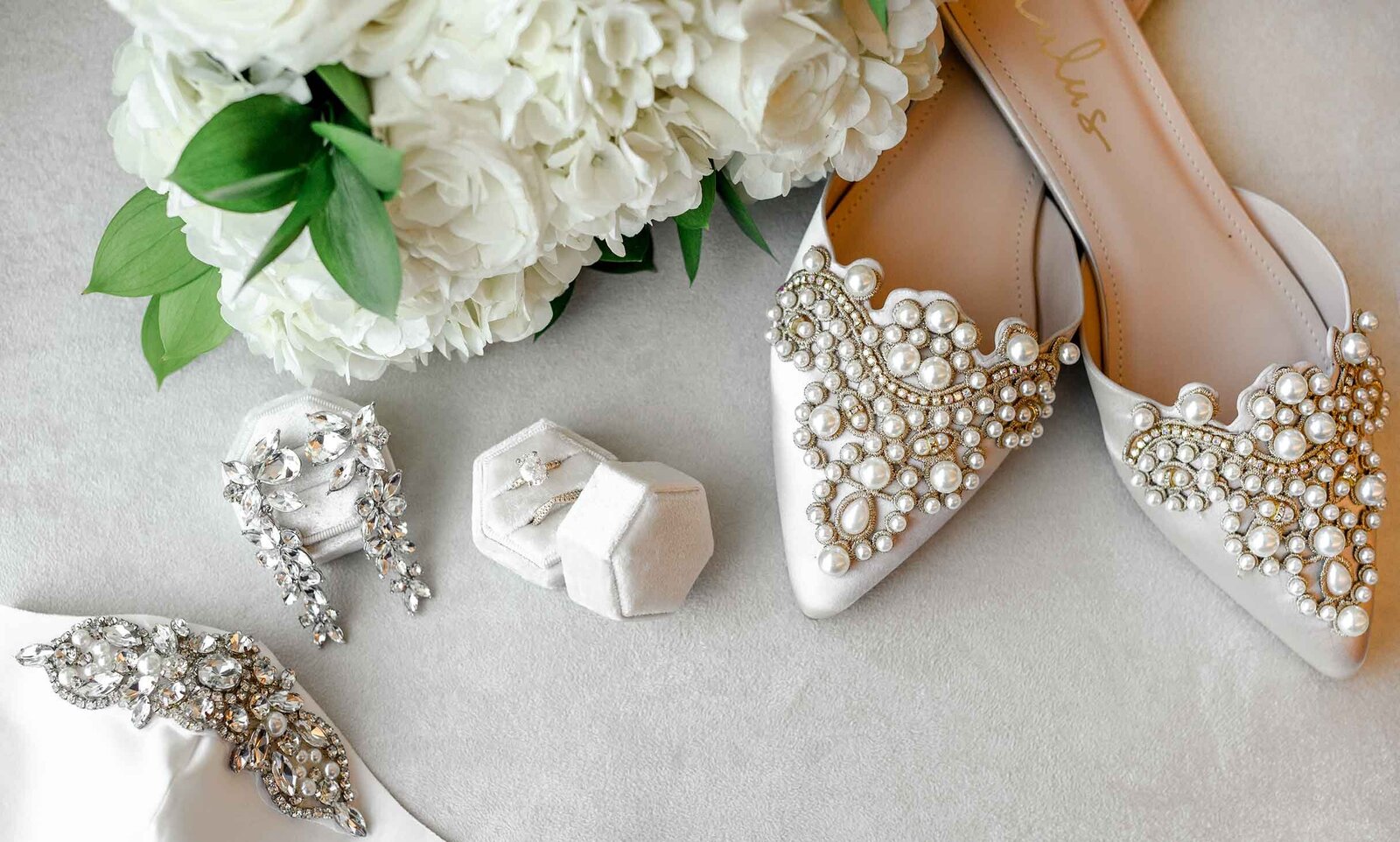Flatlay of white wedding details including flower bouquet, ring, and jewelry