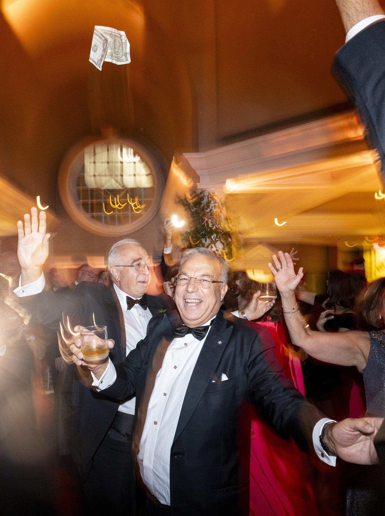 A joyful man in a tuxedo dances energetically at an event, holding a drink. Other well-dressed attendees surround him. A dollar bill is floating in the air above the crowd, captured in motion against a warmly lit venue.