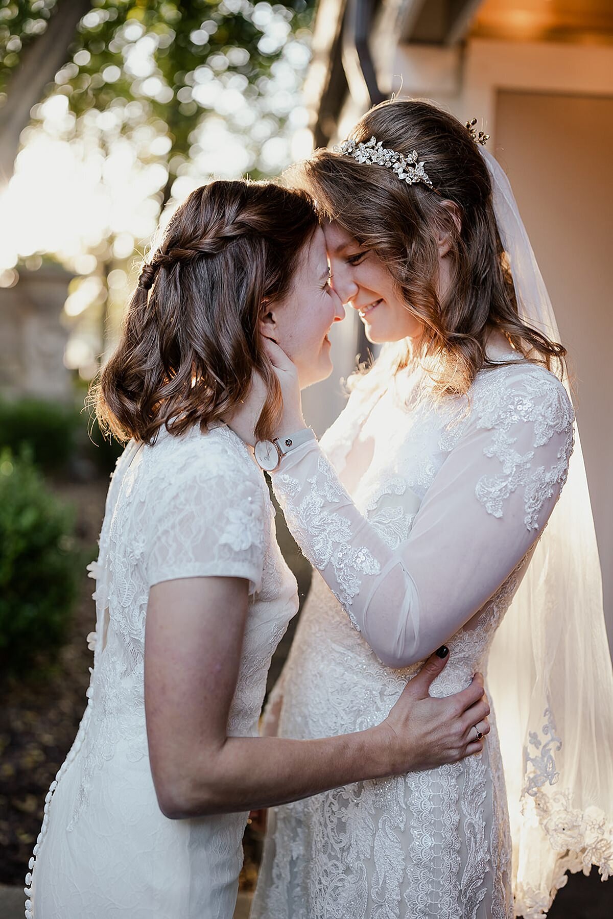 Tall bride wearing a white lace dress, veil and tiara kisses her wife, another bride wearing a short sleeved white lace wedding gown