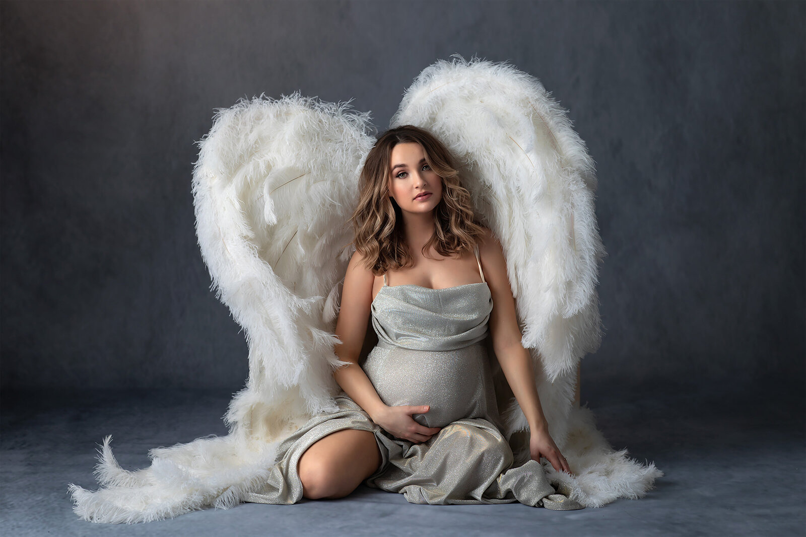 Pregnant woman sits gracefully in a shimmering silver gown with large white angel wings framing her. She cradles her belly against a soft gray backdrop in this ethereal fine art portrait by DFW Maternity Photographer Lindsay Walden.