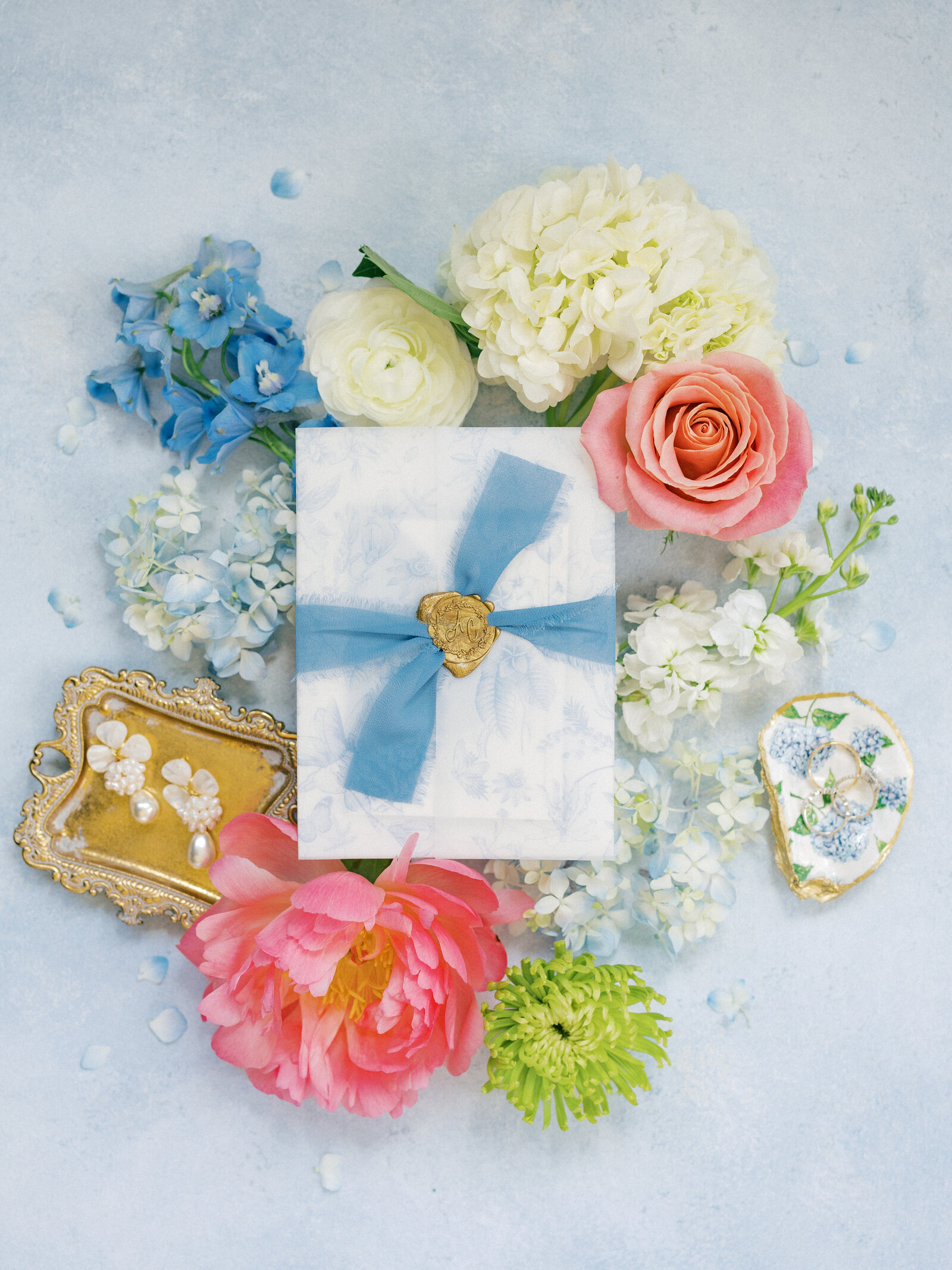 Wedding invitation with gold wax seal and blue ribbon sits with colorful flowers surrounding it