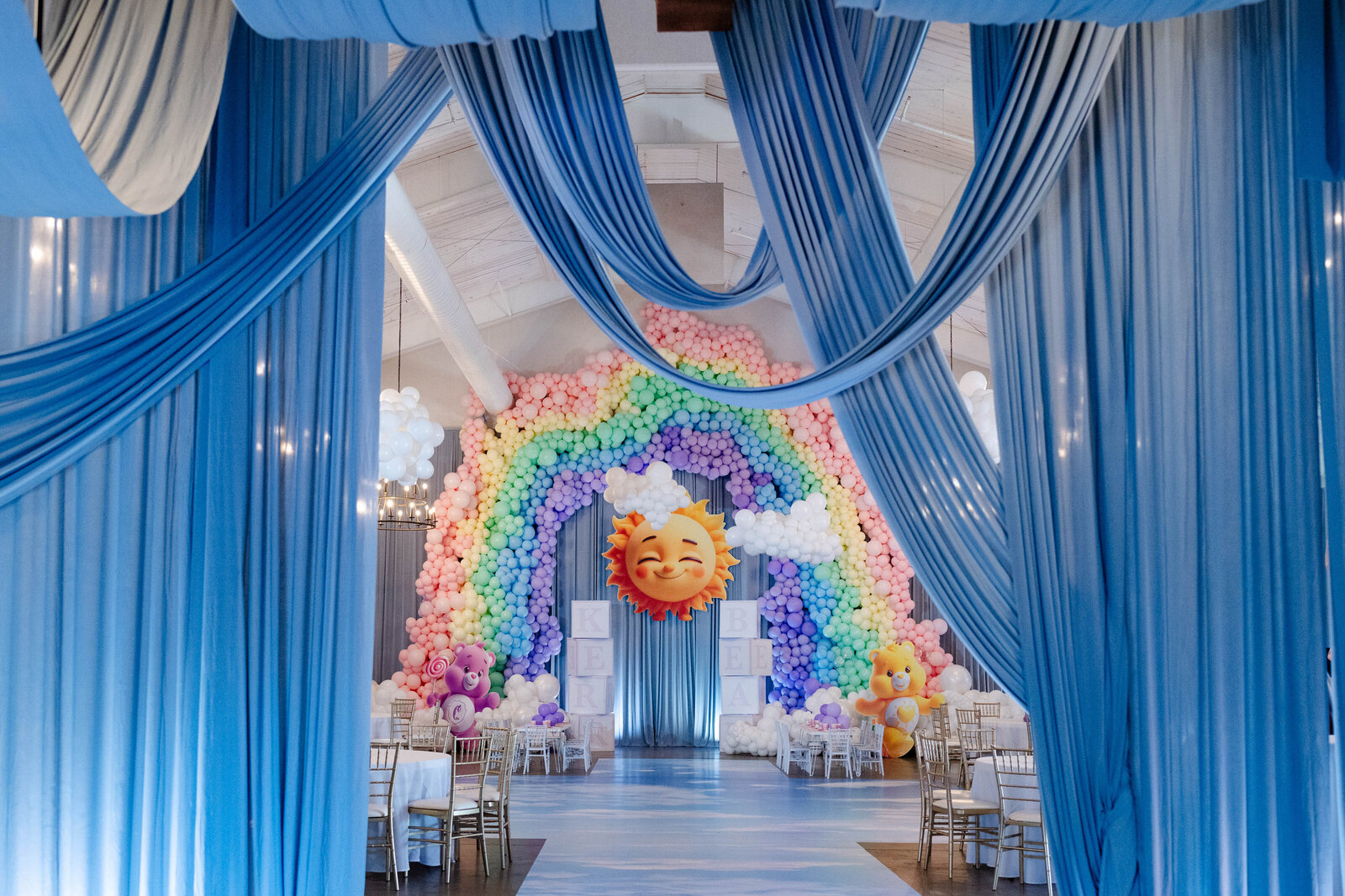 Pastel Care Bears-themed birthday party with rainbow balloon arch, blue drapery, smiling sun backdrop, and luxury event design by Wells Design Co