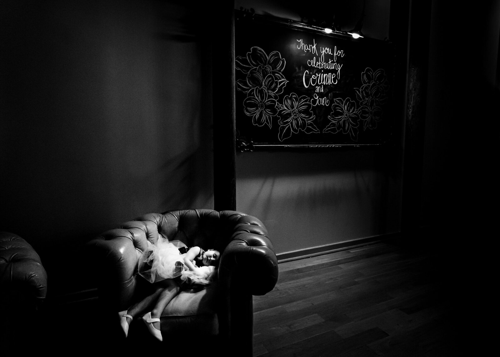 flower girl is pictured asleep in a chair in front of a chalkboard welcoming guests to the wedding reception