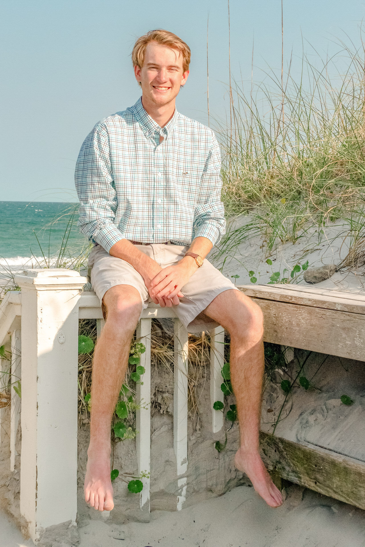 A high school senior in a checkered shirt and shorts sitting on a beach fence with the ocean and dunes behind him in Wrightsville Beach, North Carolina. This relaxed image exemplifies casual and natural senior photography, perfect for capturing youthful spirit.