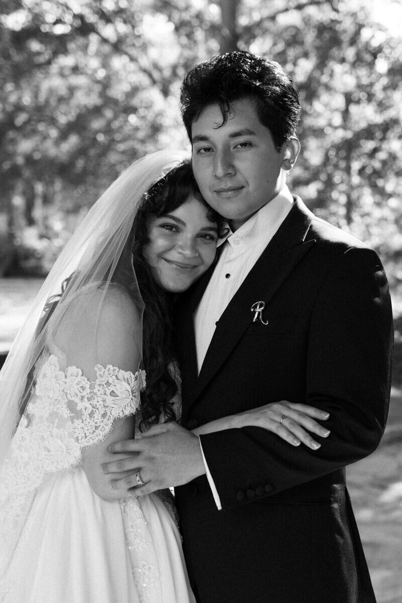 A bride and groom stand closely together, smiling softly. The bride wears a lace off-shoulder dress and veil, while the groom is in a dark suit with a boutonniere. They are outdoors with blurred greenery in the background. Black and white image.