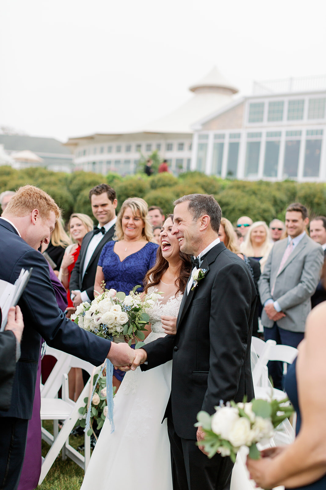 Man shaking a grooms hand as a bride laughs next to them
