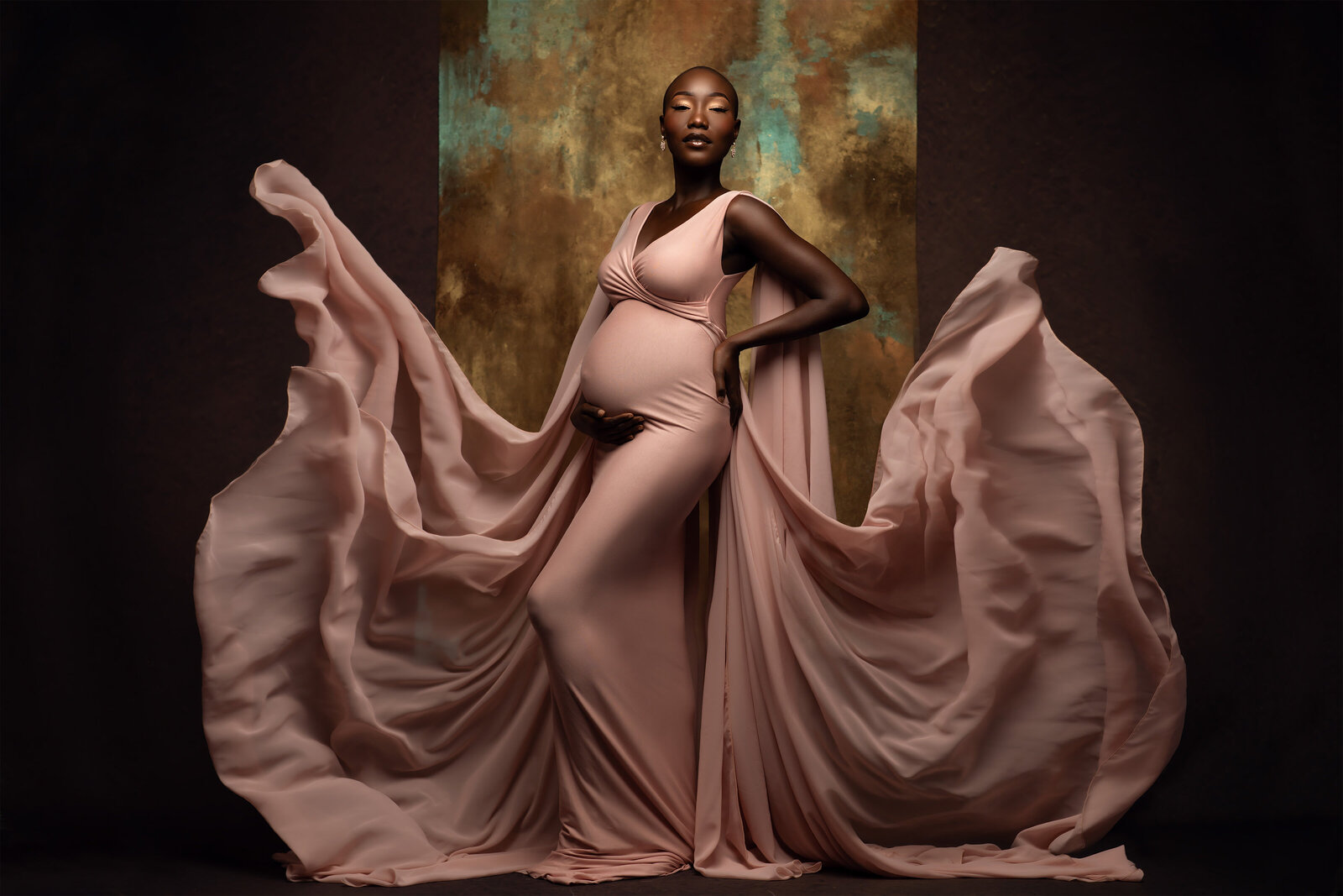 A stunning pregnant woman poses gracefully in a blush pink gown with dramatic flowing fabric surrounding her. Her hand rests on her belly as she stands against a gold and teal textured backdrop. Photo by DFW Maternity Photographer Lindsay Walden.