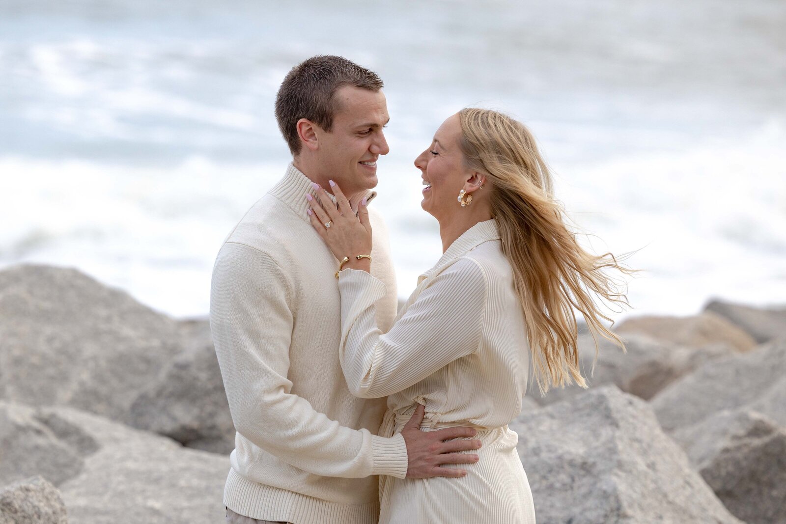A joyful couple laughs together on rocky a Fort Fisher NC beach, dressed in matching cream outfits that complement the natural tones of the ocean and rocks. The breeze blows softly through the woman’s hair, adding a touch of movement to this vibrant engagement photography moment.  
