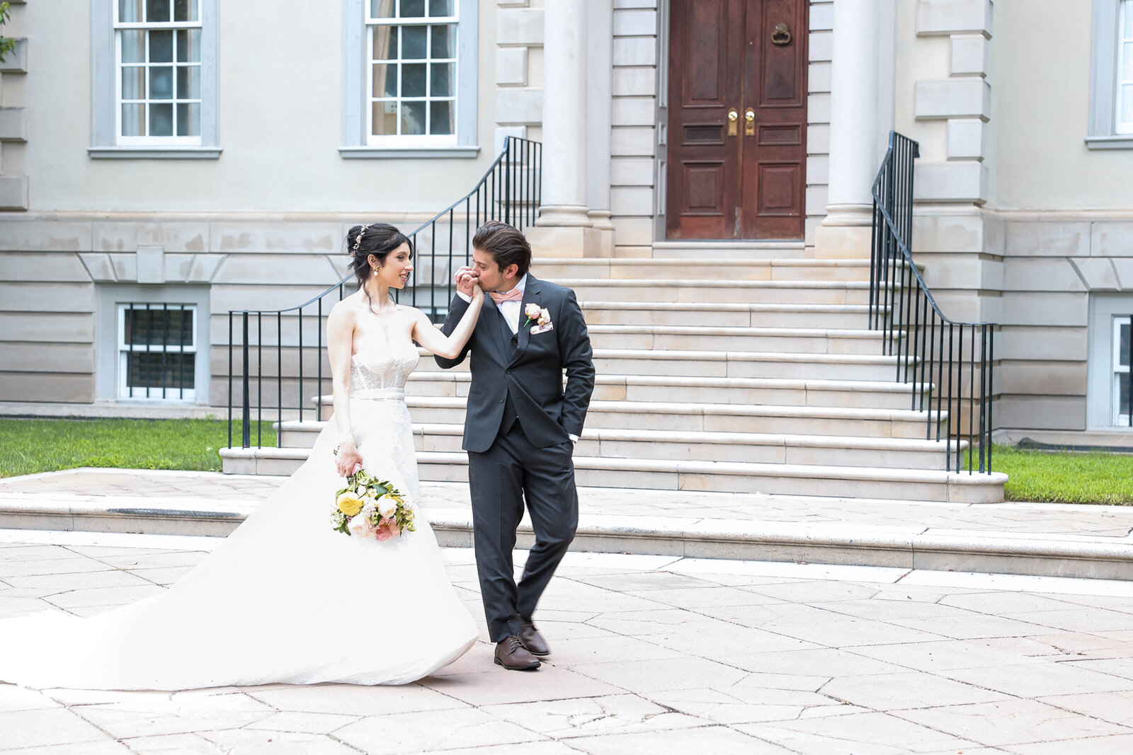 A bride and groom share a tender moment in front of a grand staircase and historic building. The bride is in a flowing white gown holding a bouquet, while the groom kisses her hand, dressed in a dark suit.