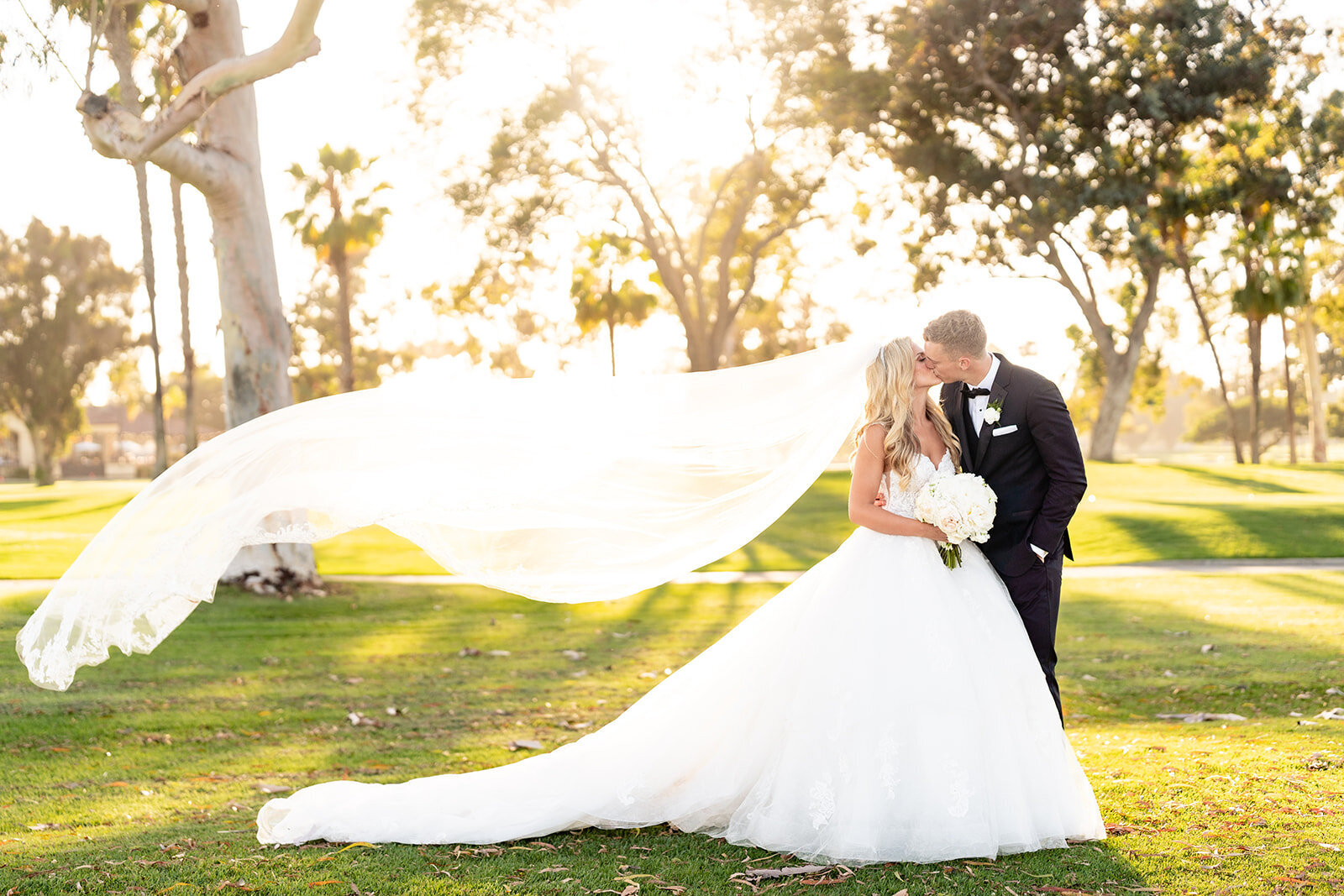 A Bride and groom kissing with her long veil blowing in the wind and the sun shining brightly through the background on a golf course at Feast and Fareway in Coronado