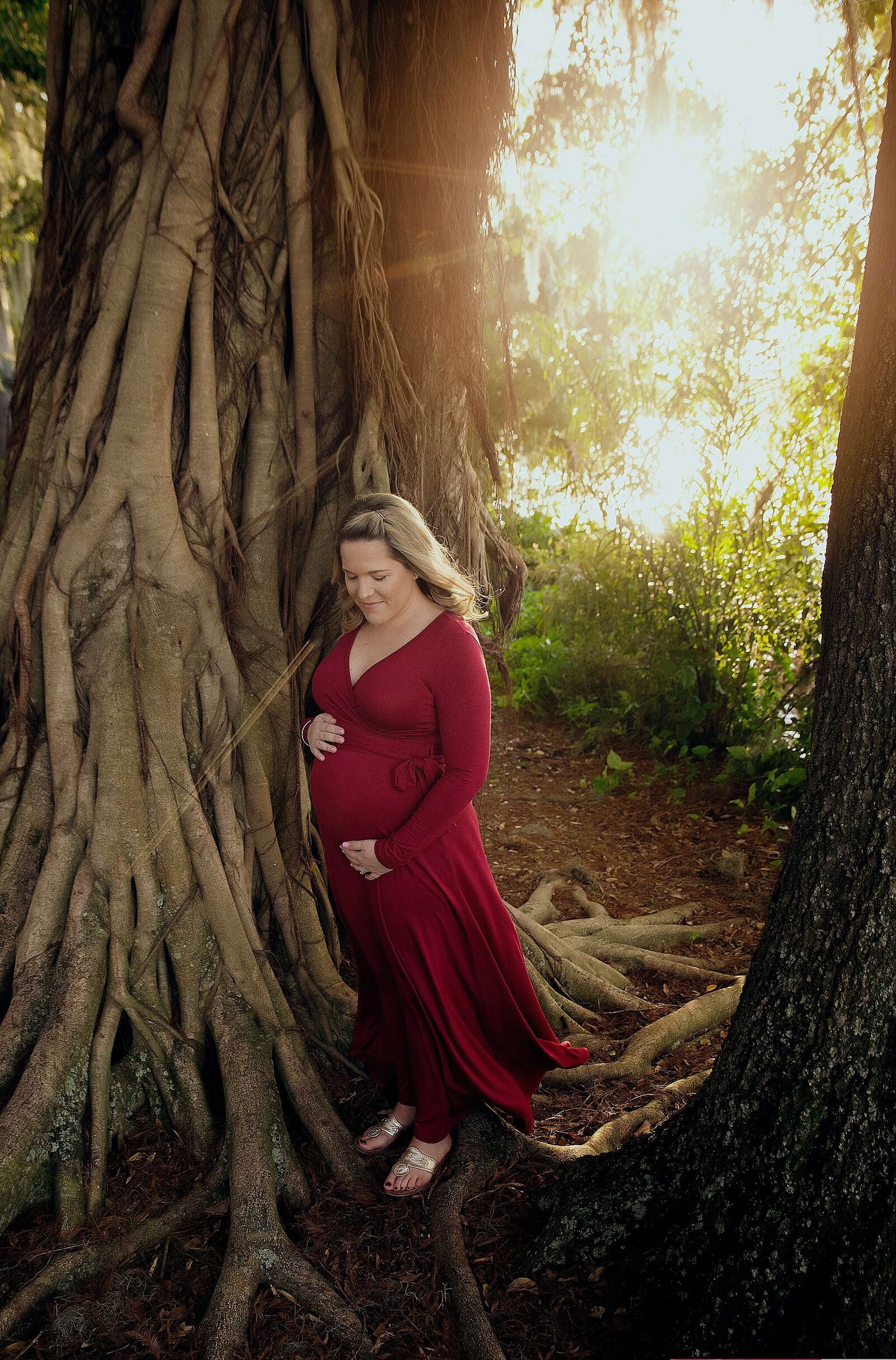 A pregnant woman in a red dress poses gracefully beside a tree, captured by Bia Schaefer, Orlando maternity photographer.
