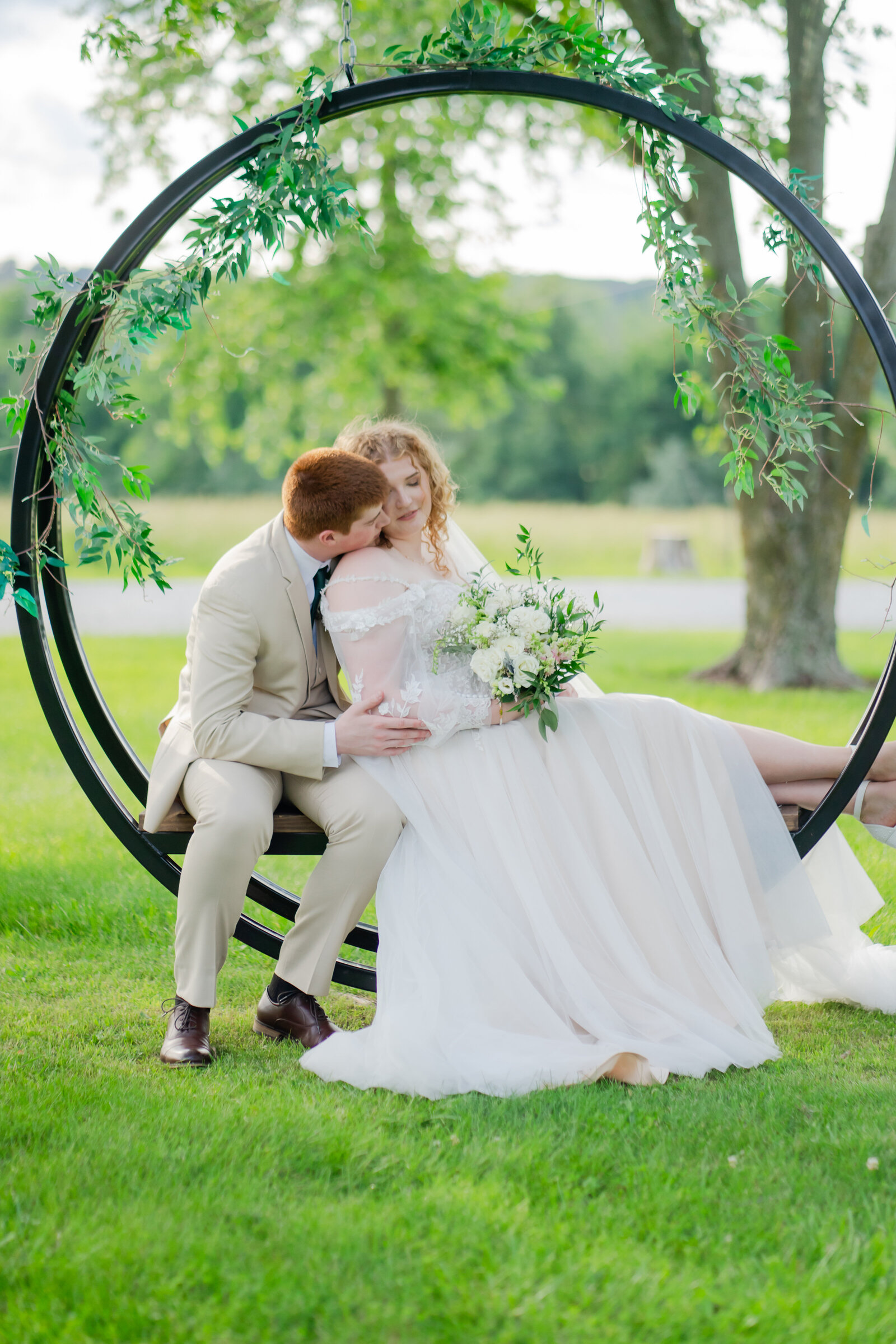 A groom wearing a tan tux is kissing the cheek of his bride. They are both sitting on a circular garden bench with an over hang. She is holding a white and green bouquet.