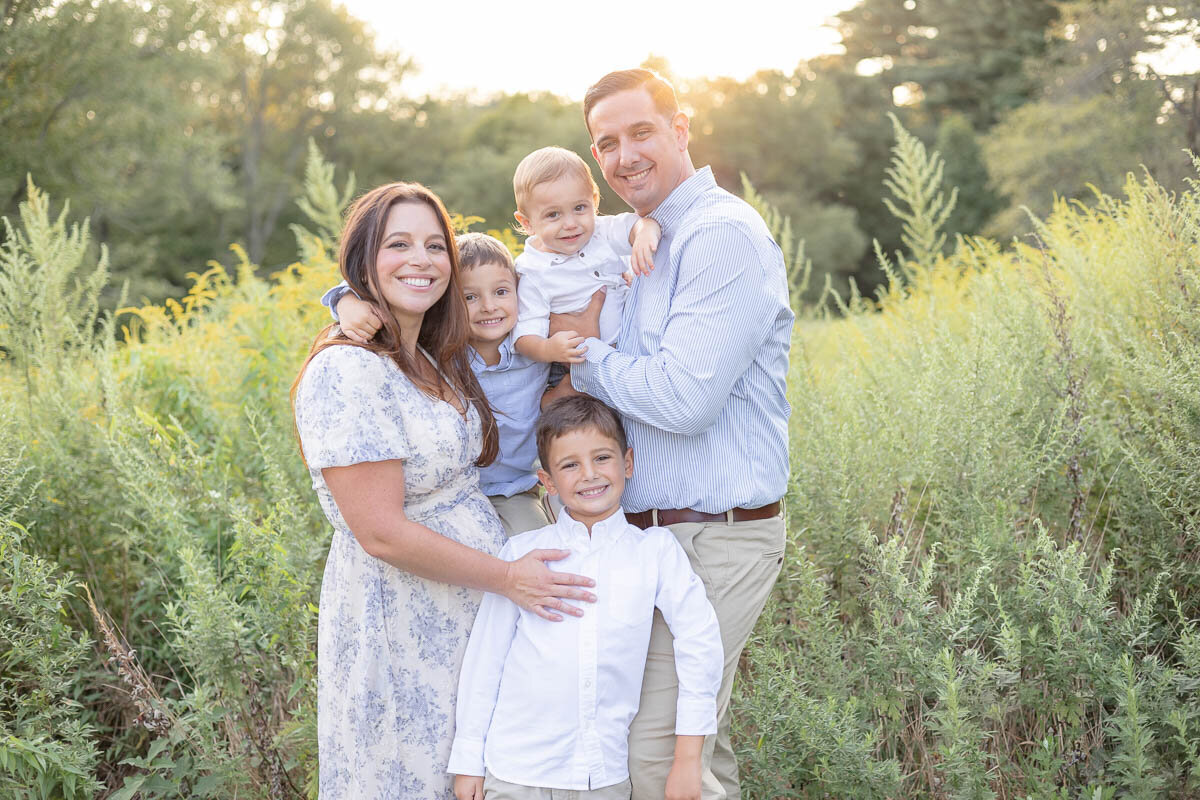 Smiling family of five poses together in a sunlit field during golden hour, captured by Westchester family photographer Nicole. Mom wears a floral dress, dad and boys wear coordinating blue and white outfits.