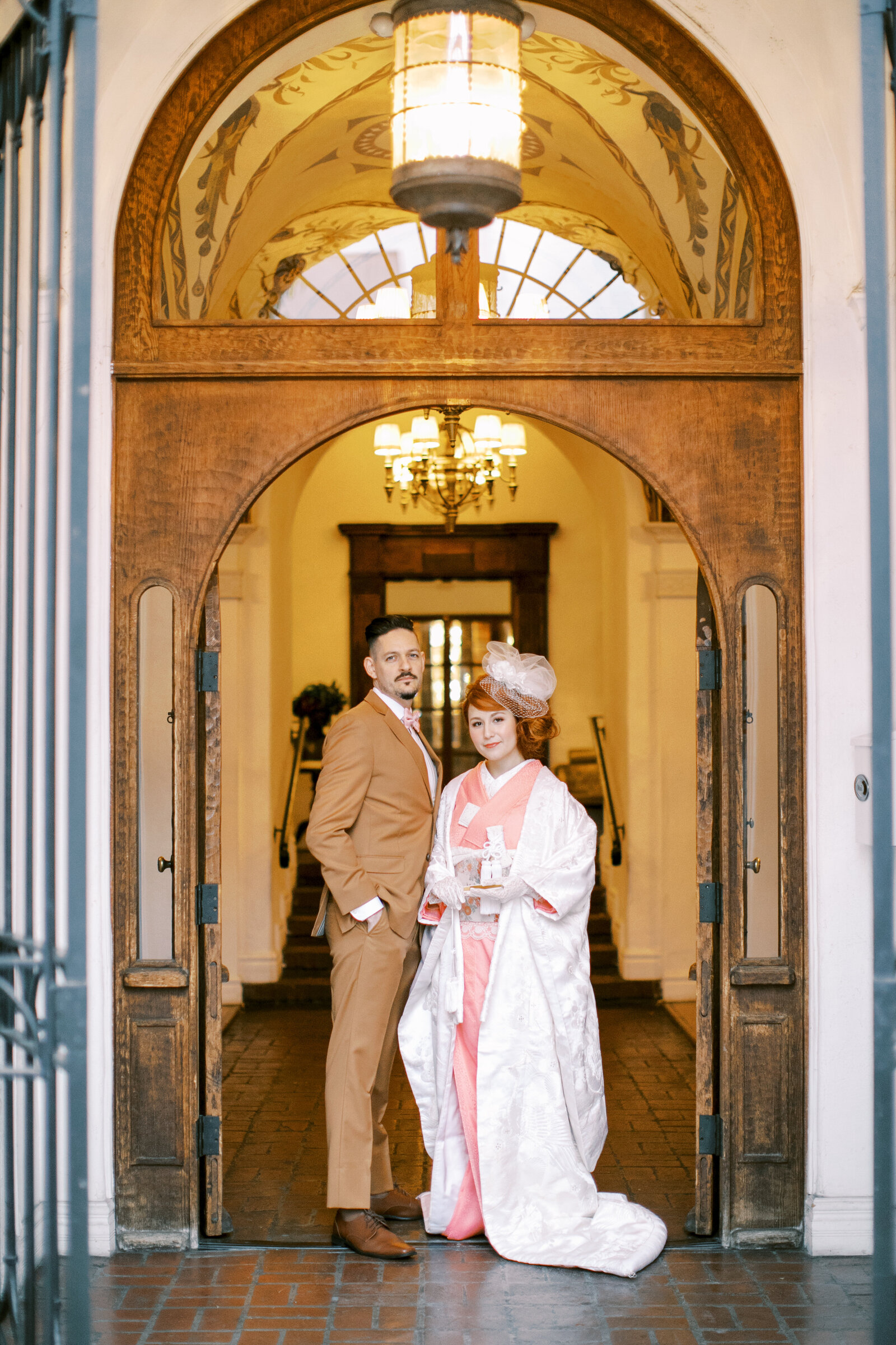 Bride and groom posing together beneath the grand arched doorway at Carondelet House, Los Angeles California. Bride wears a white shiromuku with soft pink accents. Available for wedding kimono rental | ロサンゼルスのカロンデレットハウス、大きなアーチの下で並ぶ白無垢と淡いピンク掛下の花嫁と新郎。着物レンタル可能。