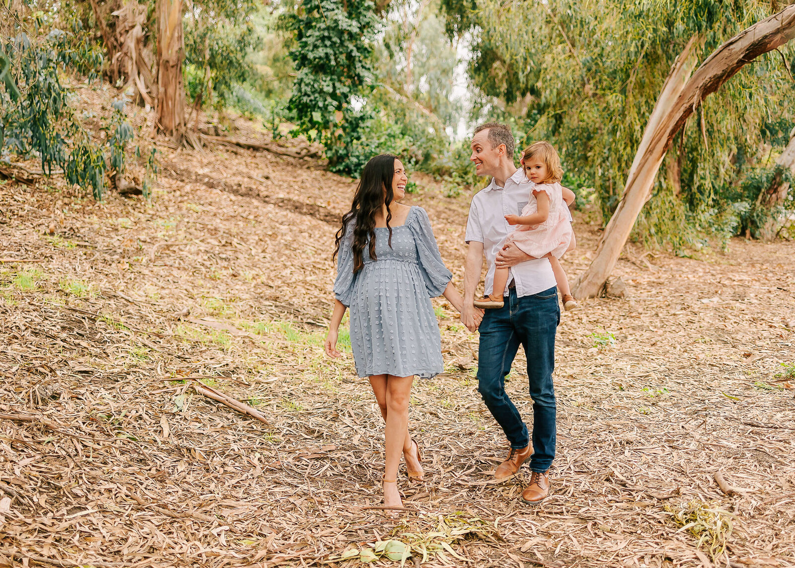Husband and wife holding hands walking with toddler on hip during during nature maternity session in Seal Beach, CA.