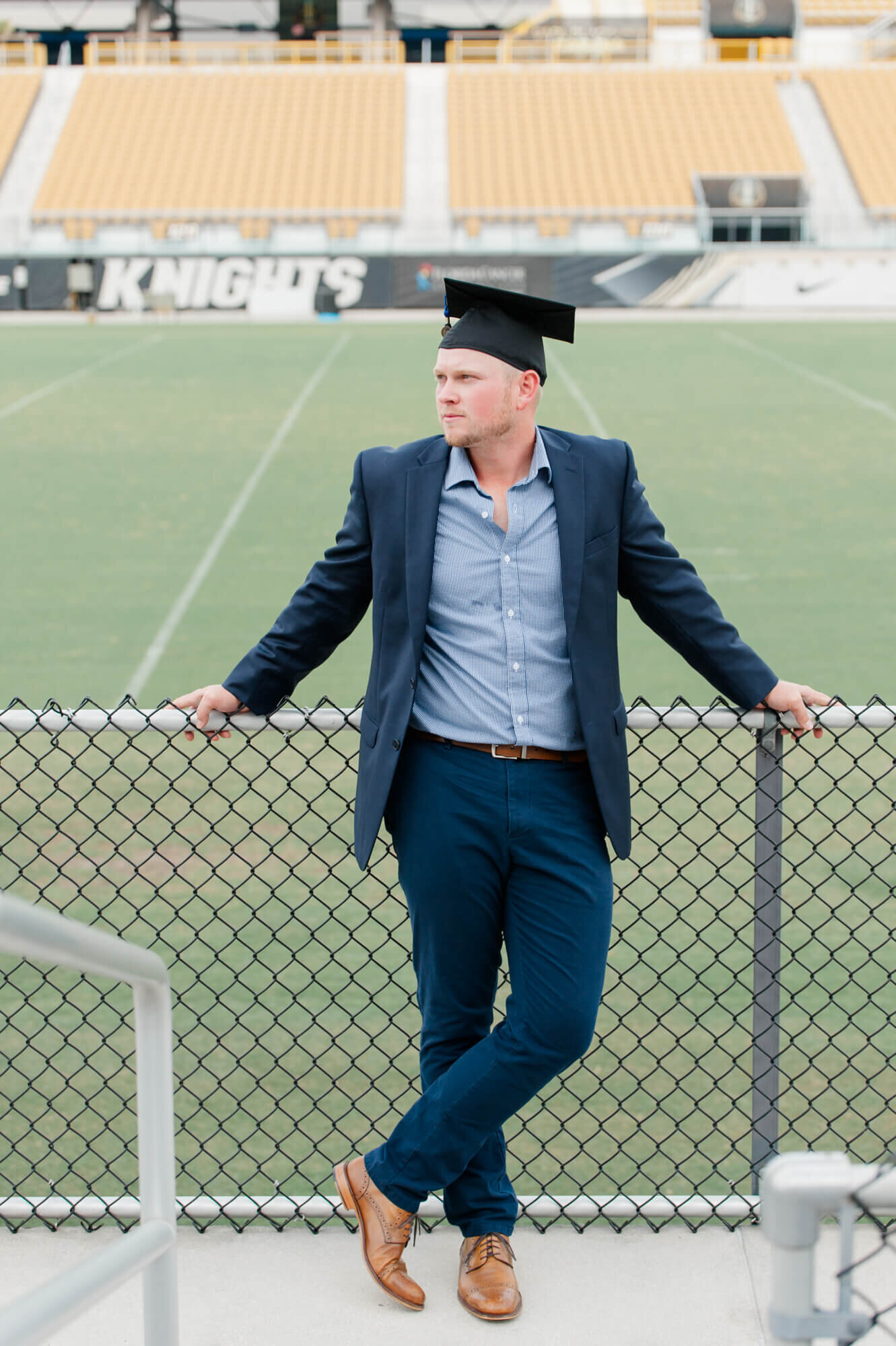 Senior male grad stands against the fence at UCF football field and reminisces on the good ol days