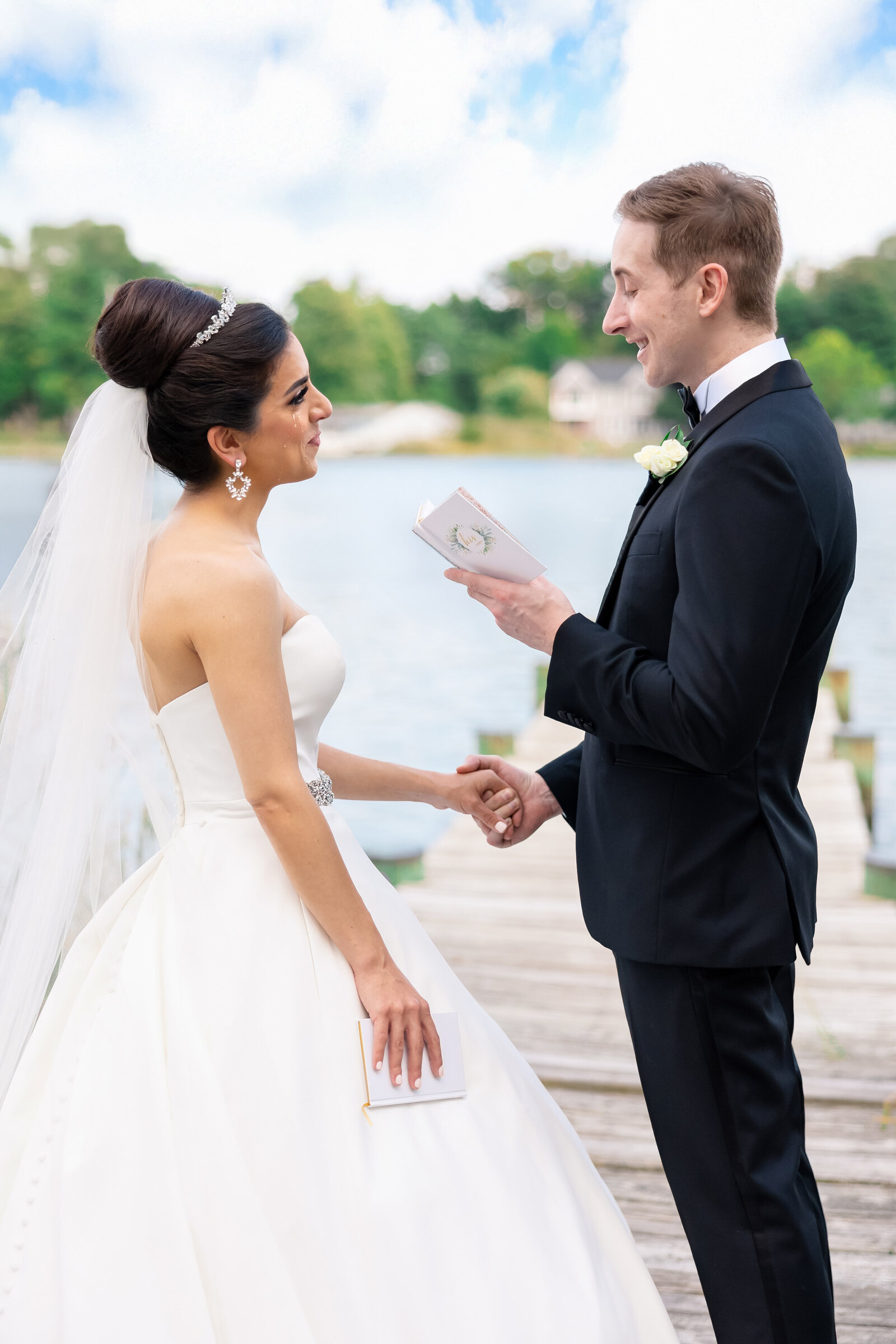 A bride and groom exchange vows on a wooden dock by a lake. The bride wears a strapless white gown and veil, holding her wedding vows. The groom is in a black suit with a white boutonniere, reading from a vow book. Trees and sky are visible in the background.
