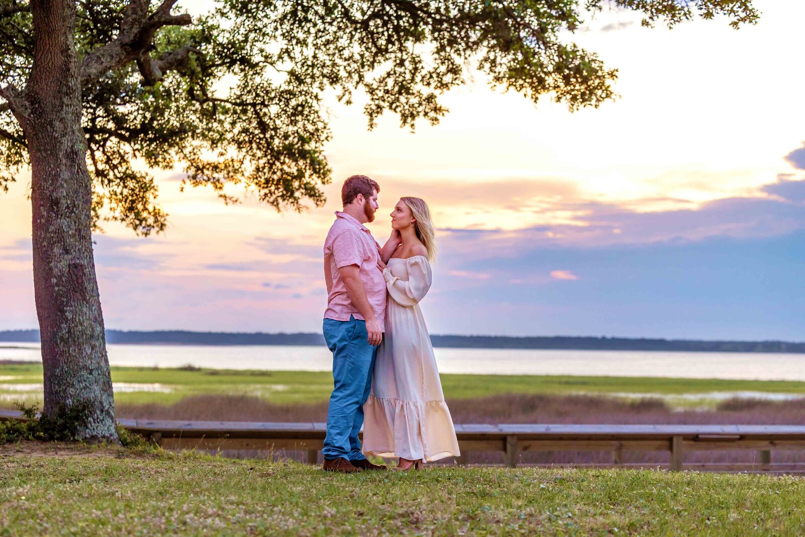 A couple stands under a tree at sunset, gazing lovingly into each other's eyes with a serene waterway and colorful sky in the background. The woman wears a flowing white dress, and the man pairs jeans with a casual pink shirt. This picturesque scene embodies the charm of North Carolina engagement photography. 