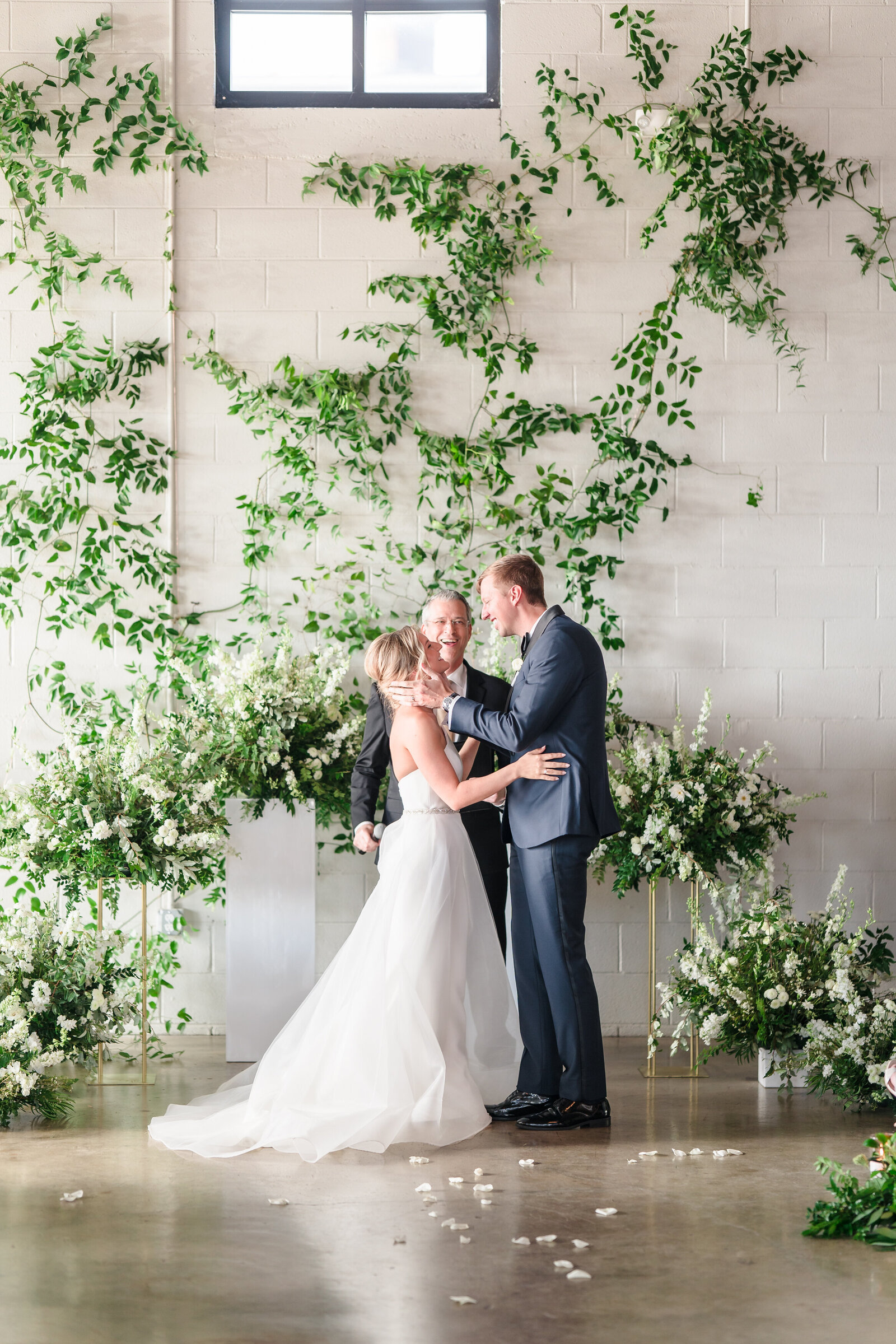 A bride and groom lean in for their first kiss surrounded by green florals with white flowers