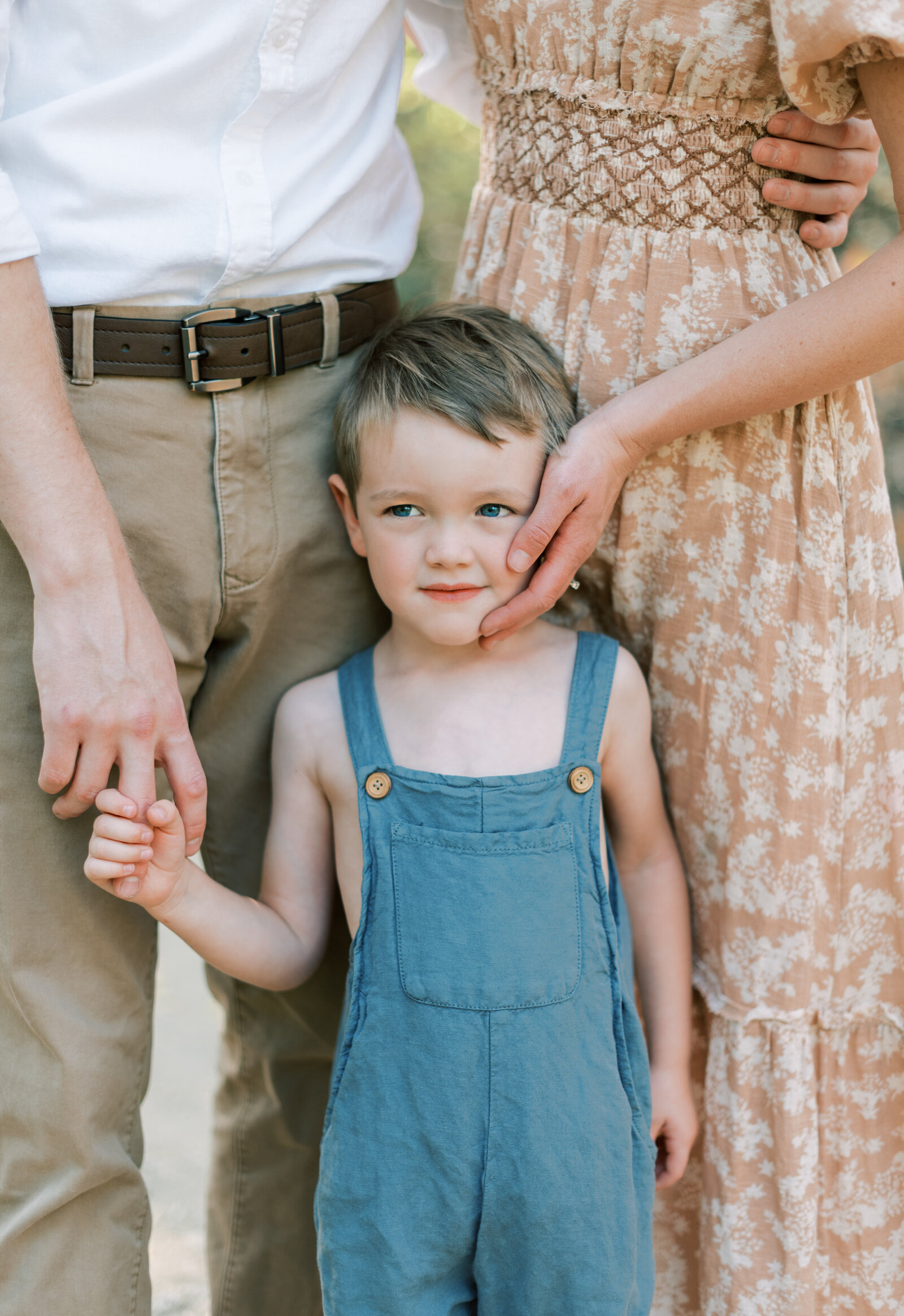 Portrait of a young boy in blue overalls nestled in between a woman in a summer dress and a man in a dress shirt and khakis