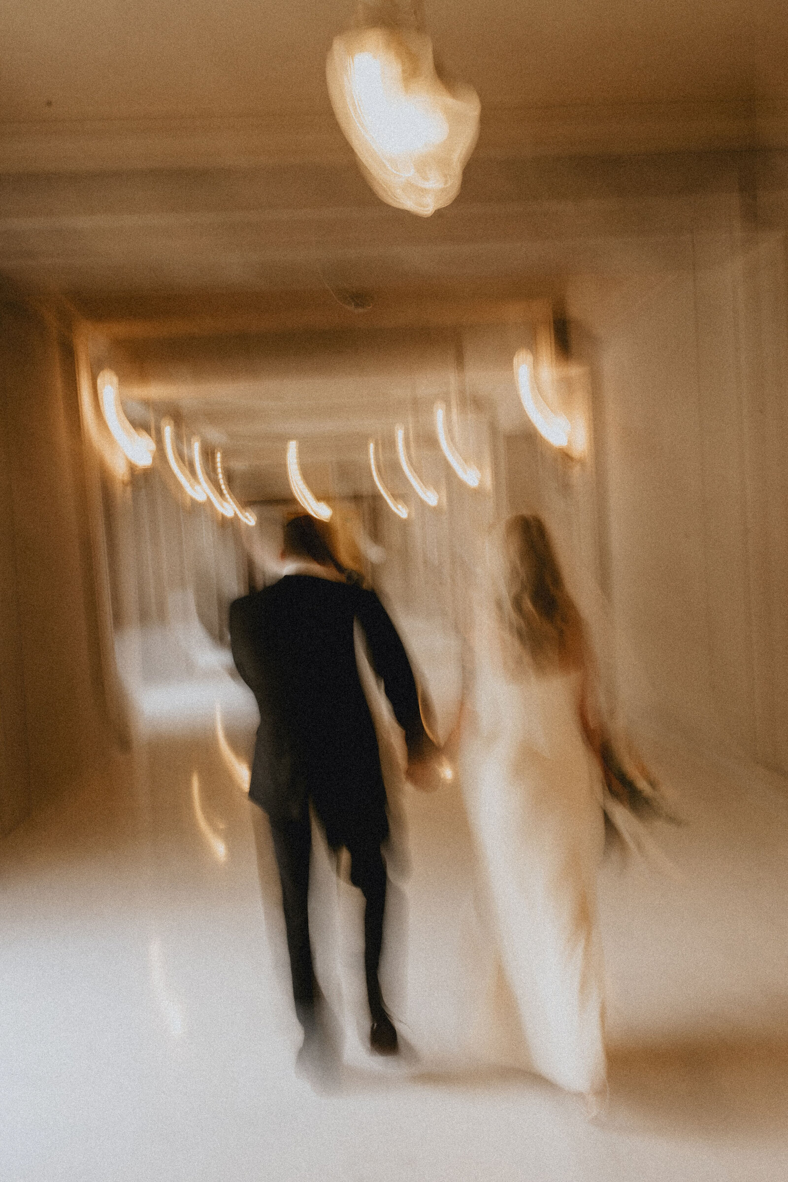 A bride and groom holding hands and walking down a warmly lit corridor in San Francisco City Hall, creating a dreamy effect with streaks of light from ceiling lamps.