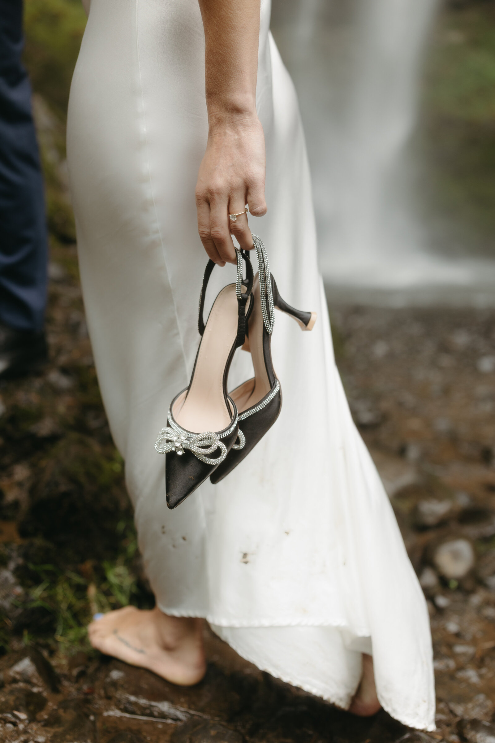 A bride is holding her wedding shoes as she climbs the rocks at a Nova Scotia waterfall.