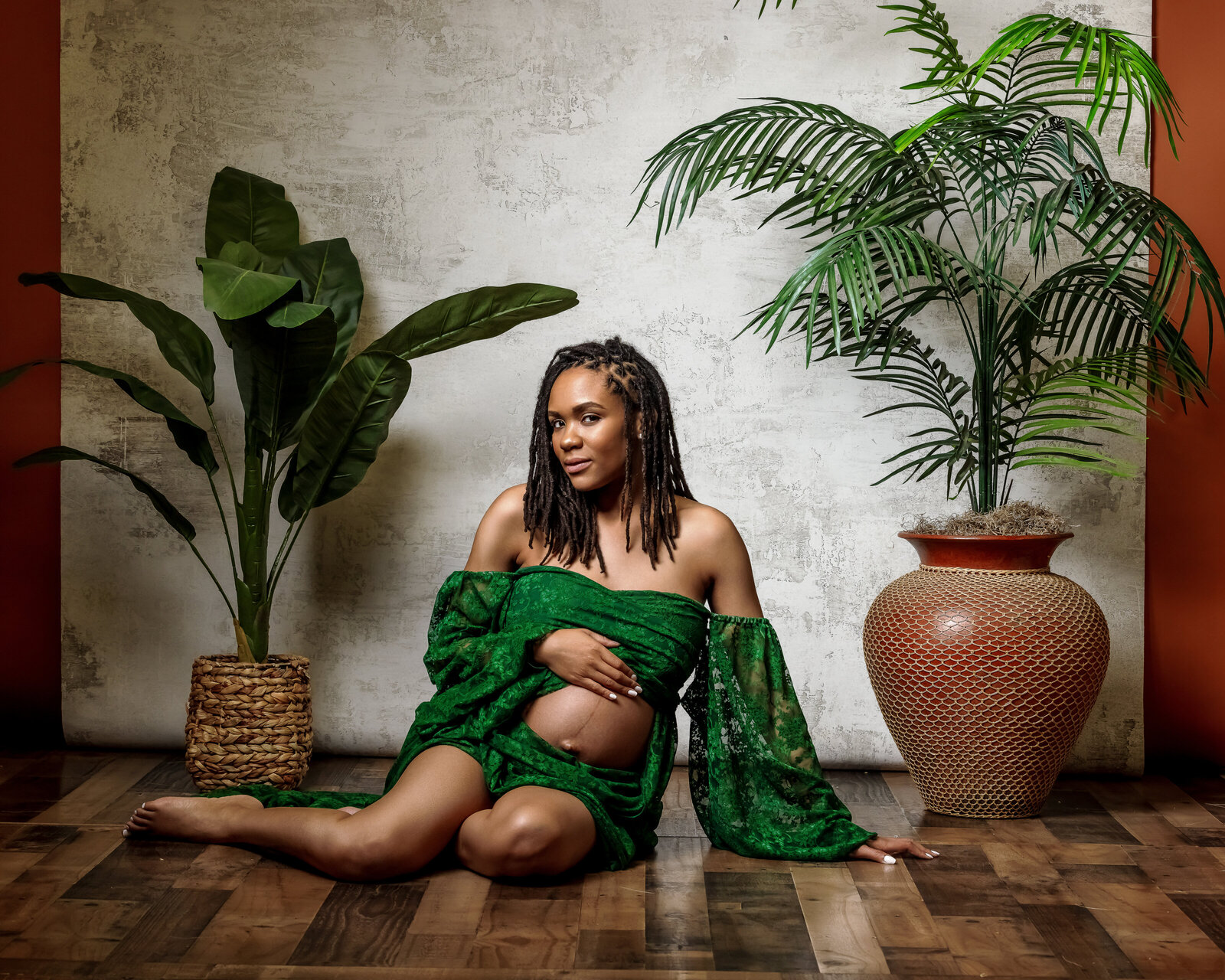African American pregnant woman sitting on the floor in a green lace gown with her hand placed on top of her pregnant stomach.