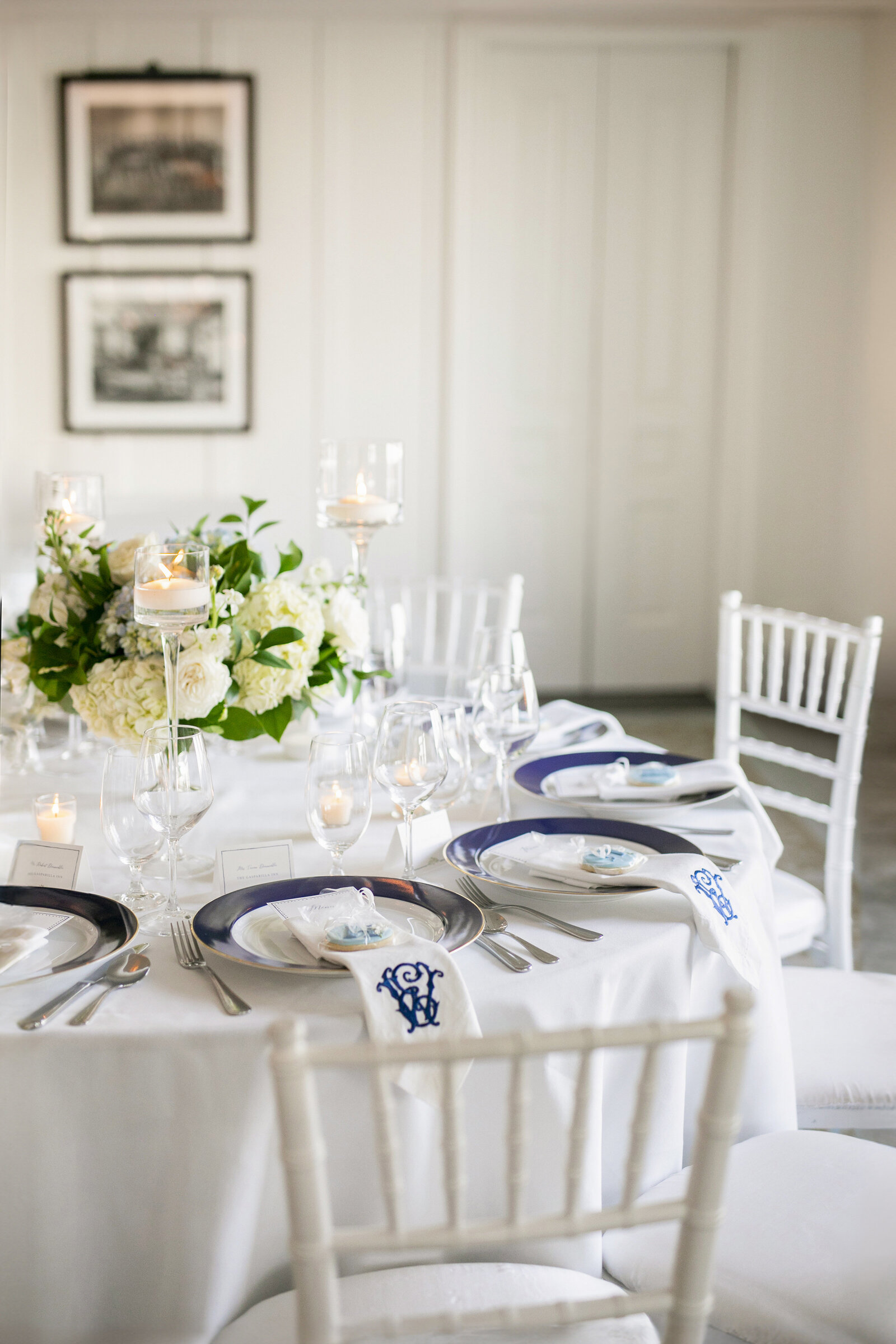 Elegant table setting with white tablecloth, silverware, blue-accented plates, and monogrammed napkins. Features white floral centerpiece, candles in glass holders, and white chairs. Black-and-white photos are on the wall in the background.