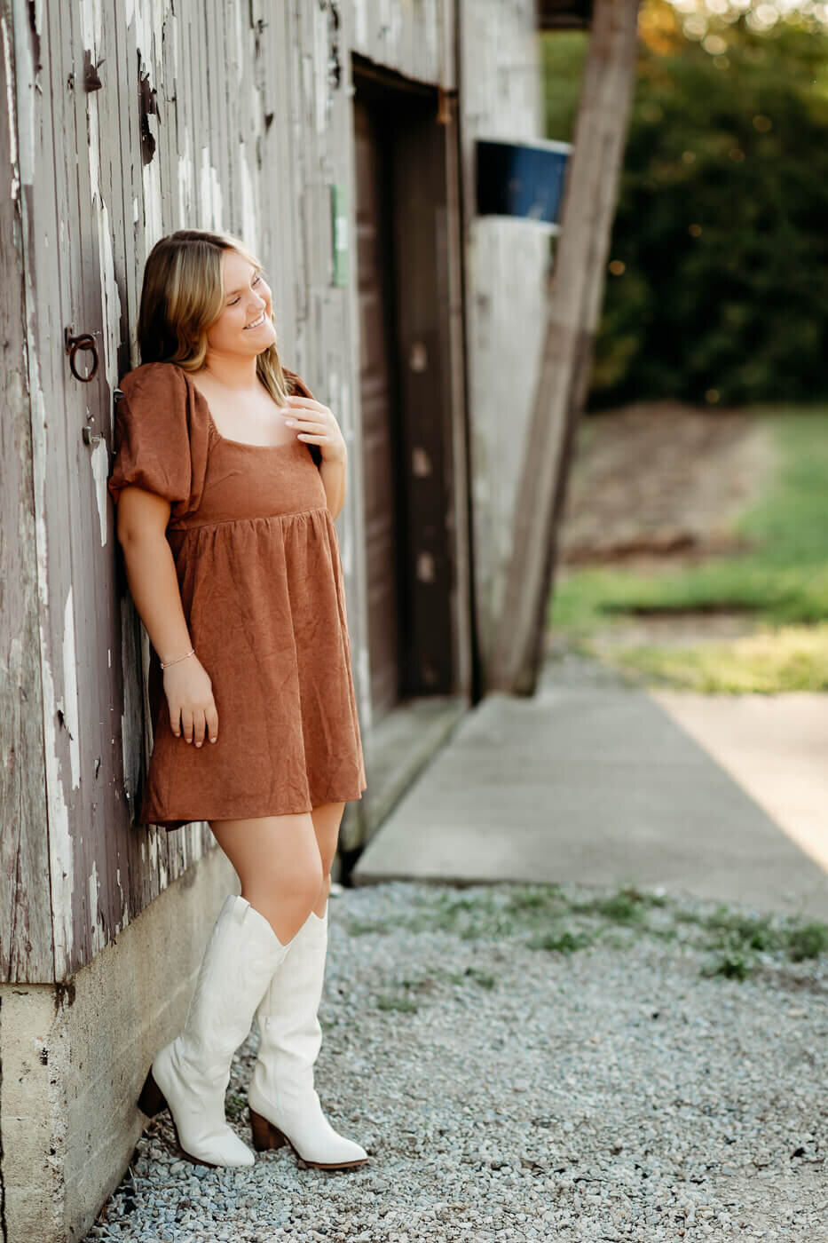 A person in a brown dress and white boots leans against a rustic wooden building, with a grassy area in the background. The sunlight creates a warm ambiance.