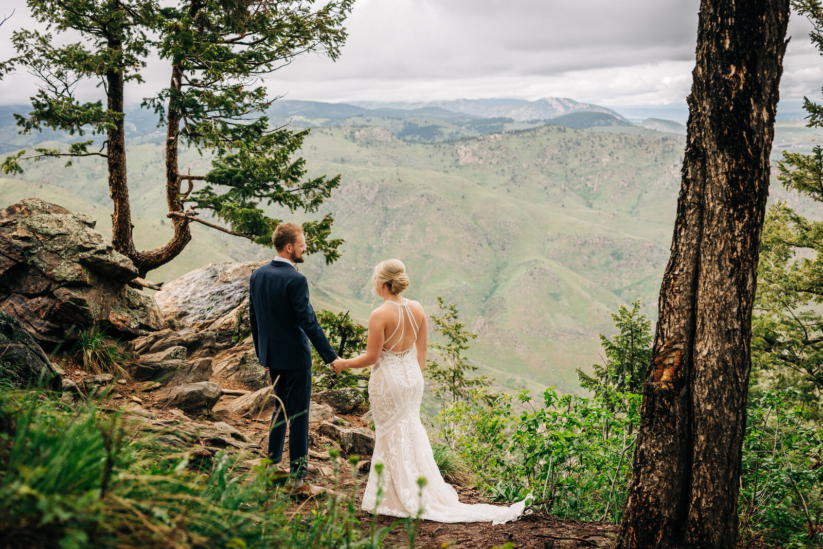 Wedding Photographer Boulder Colorado captures bride and groom holding hands and overlooking mountains in forest