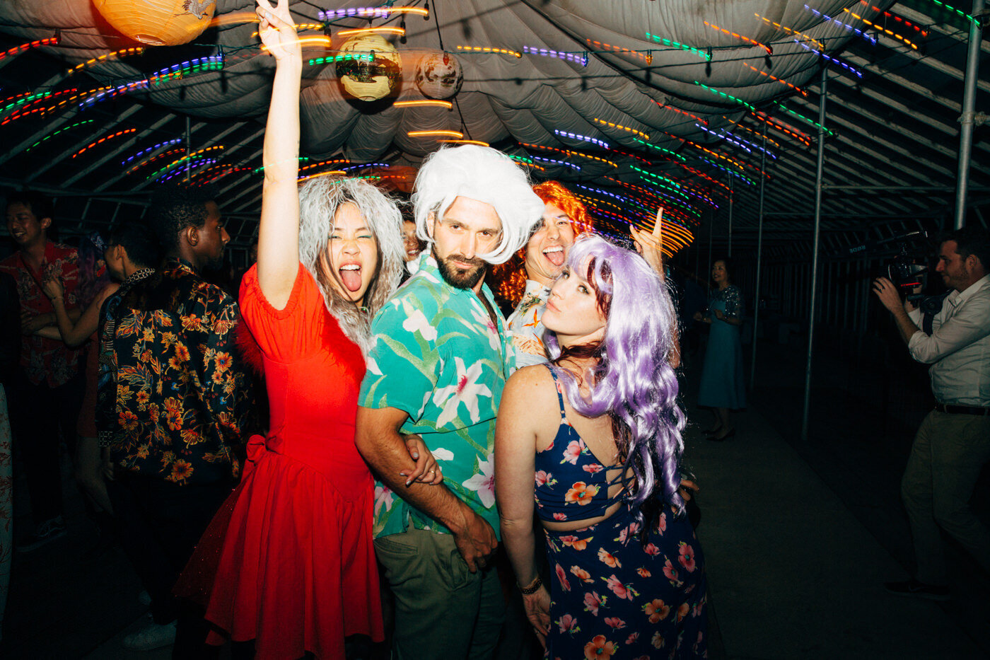 The bride and wedding guests wearing fun and colorful wigs having fun dancing at the reception under the colorful lights. Shutter drag is used by wedding photographer Megan Lynn of My Sun and Stars Co.