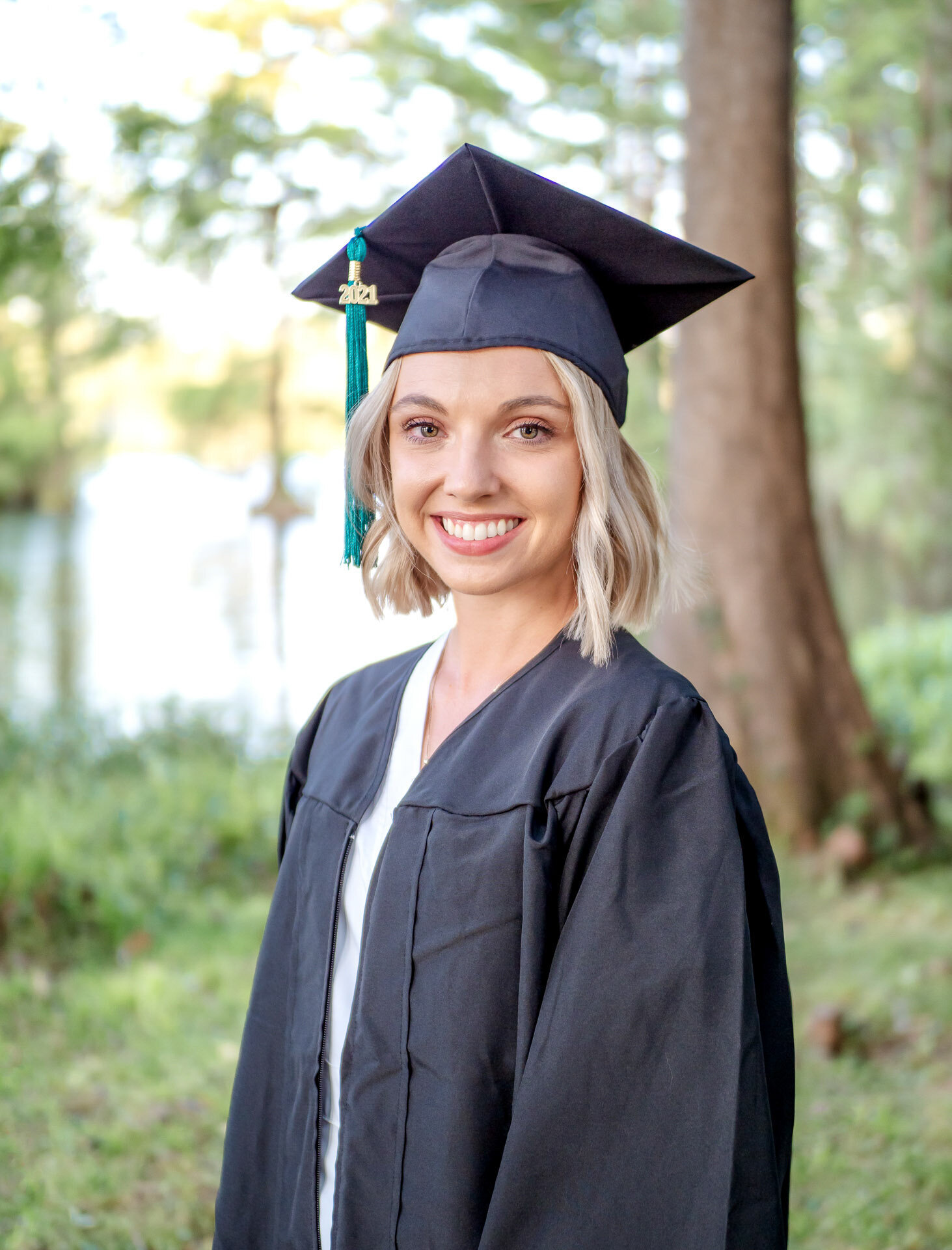A college graduating senior in a cap and gown smiling by a lakeside in Wilmington, NC. This portrait captures the essence of professional senior photography, ideal for those seeking natural and serene graduation photos.