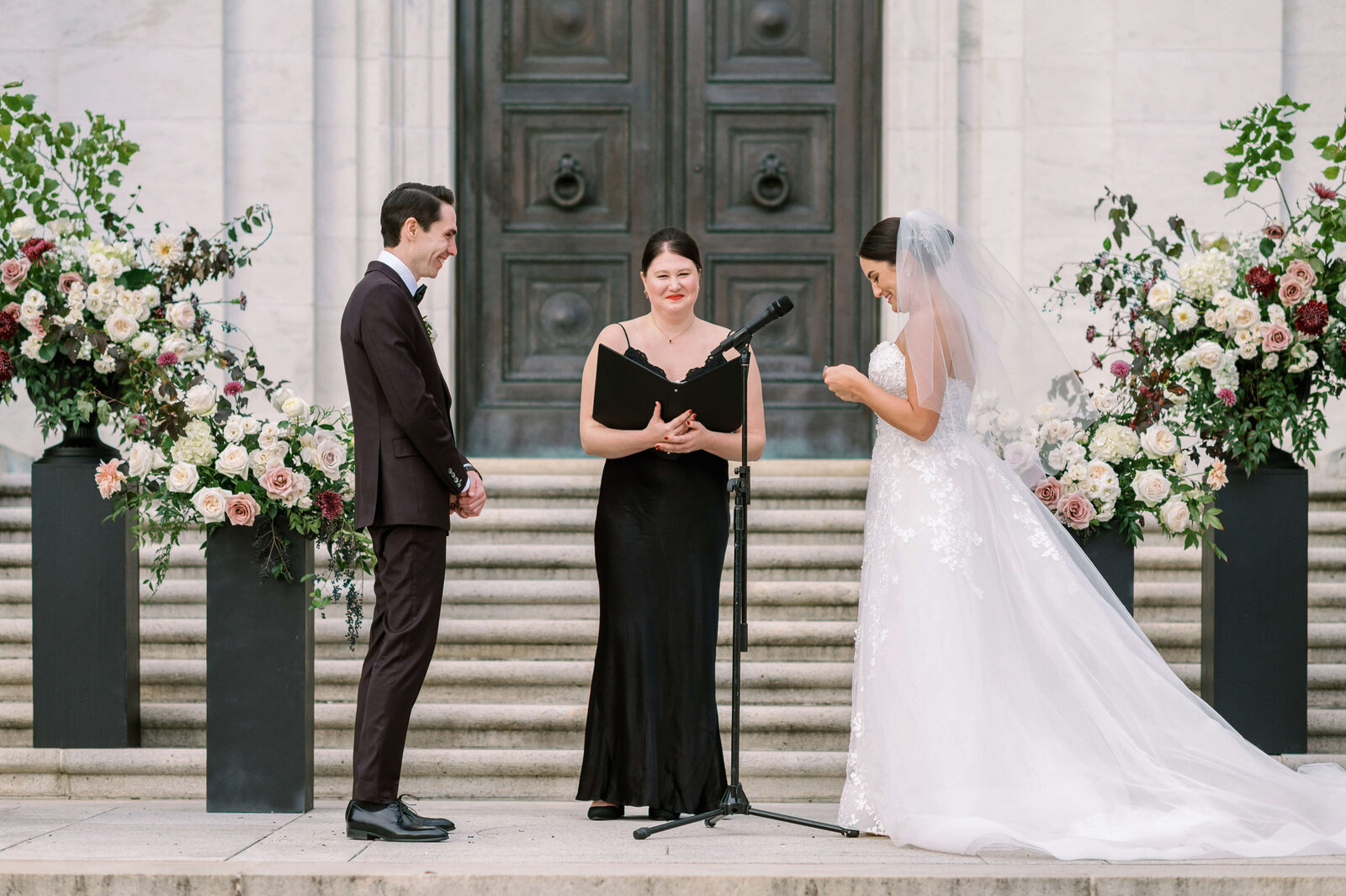 Bride in a long white wedding gown stands at the alter and reads her vows to her groom while surrounded by white and burgundy flowers